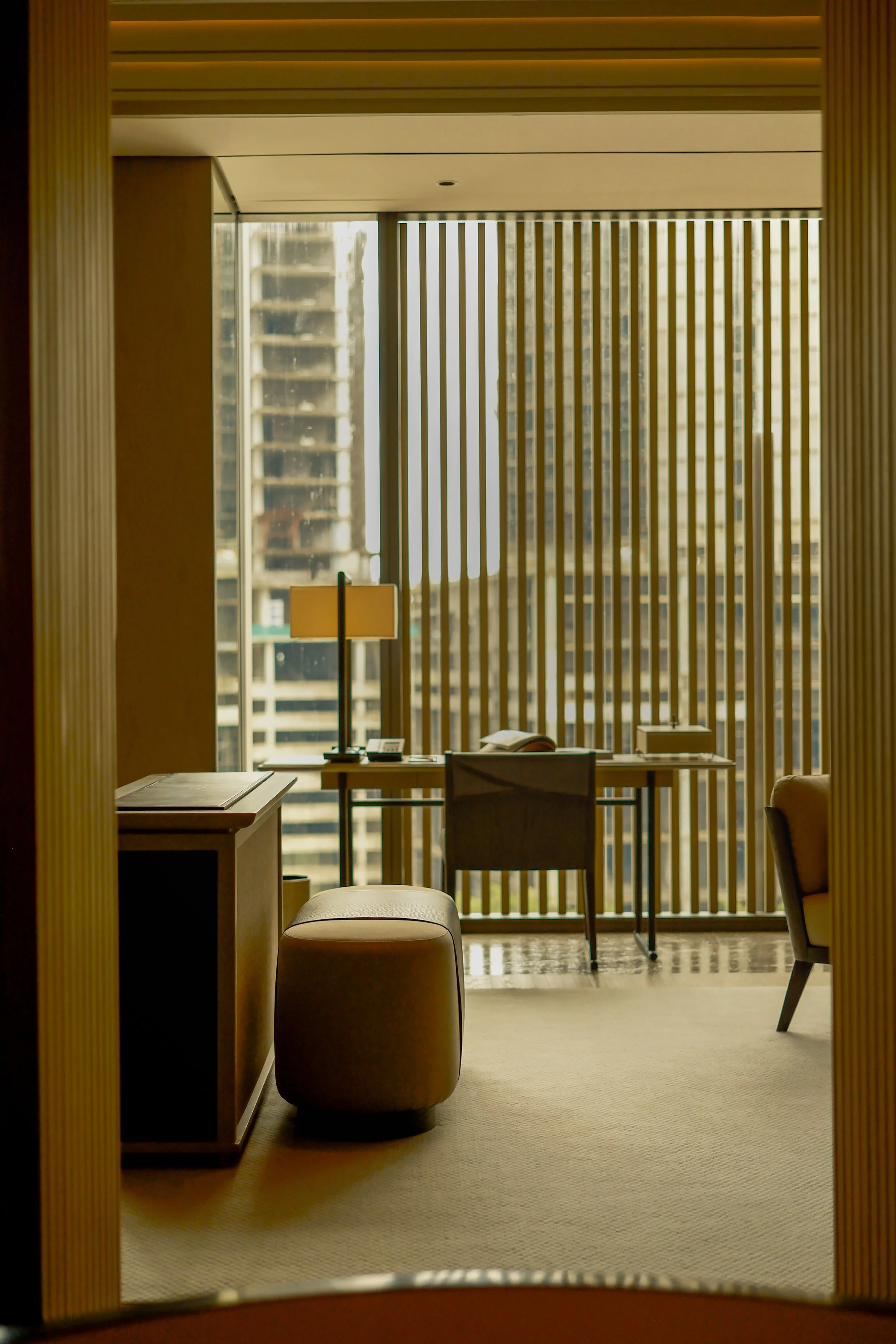View of a modern hotel room with a desk, chair, couch, and large window with vertical blinds, city buildings outside.