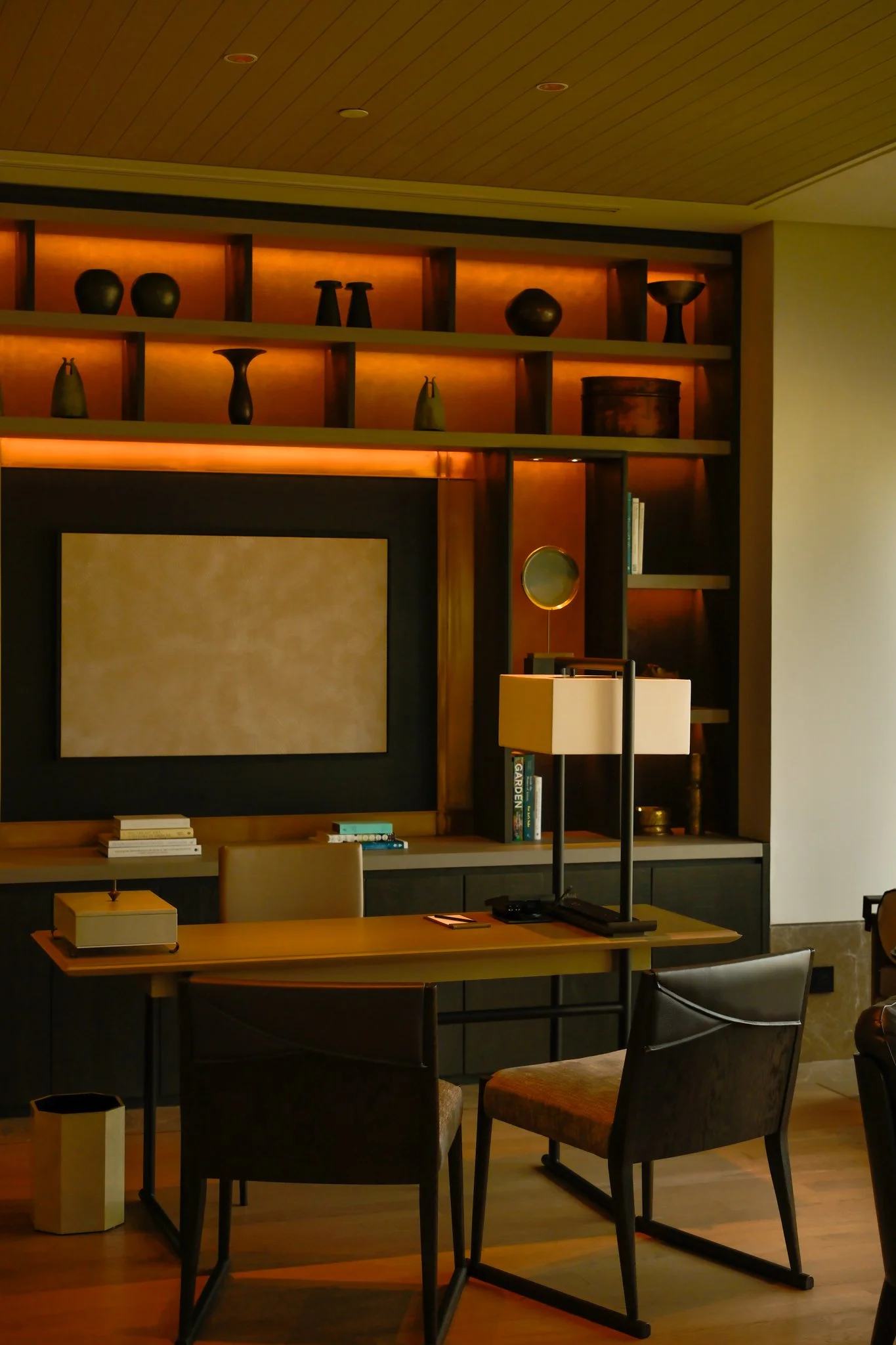 Interior of a modern living space featuring a wooden shelving unit with decorative vases and books, a table lamp, and a table with chairs.