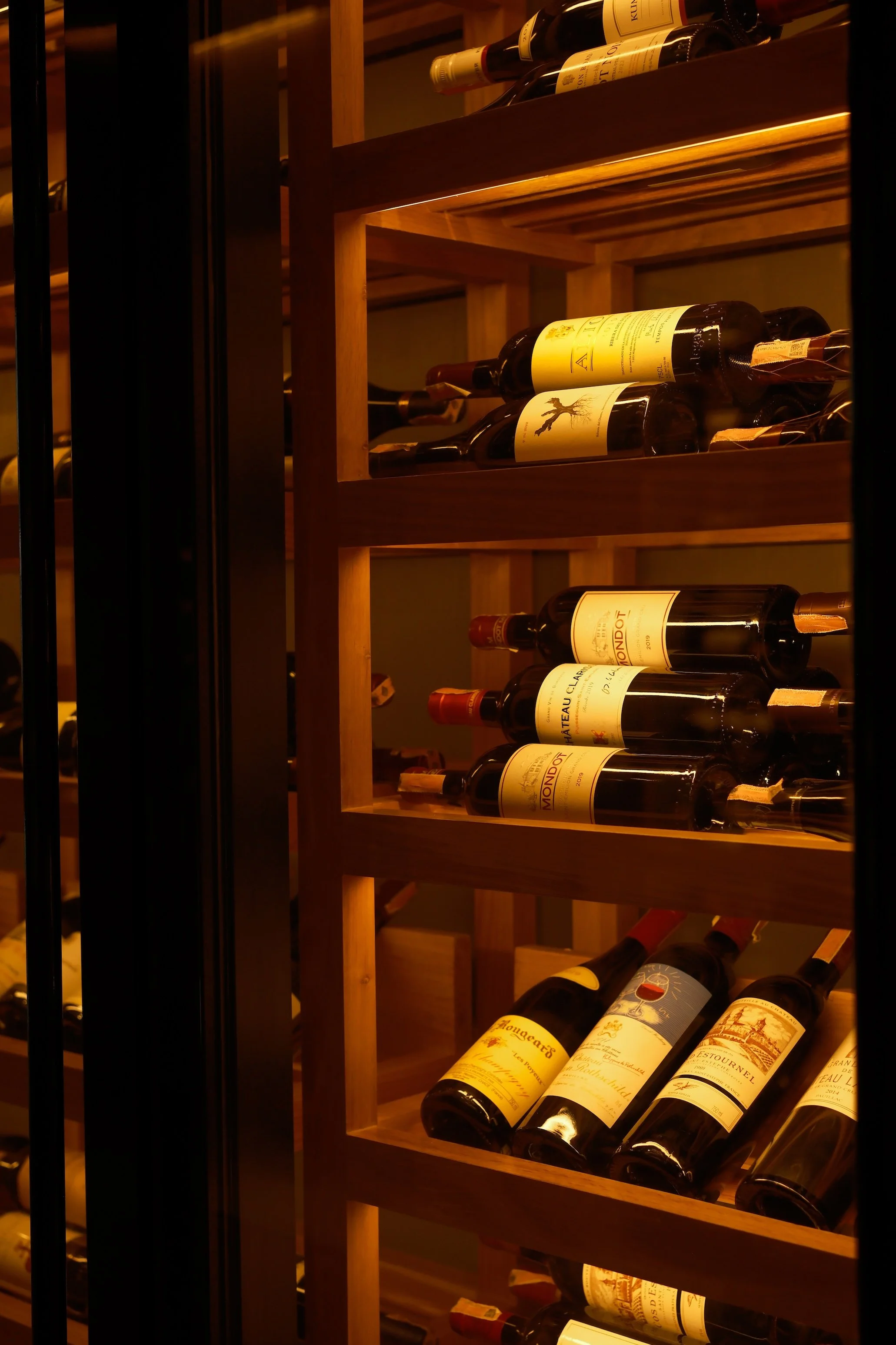 Wine bottles stored on wooden shelves inside a wine cellar or wine rack, dimly lit with warm lighting.