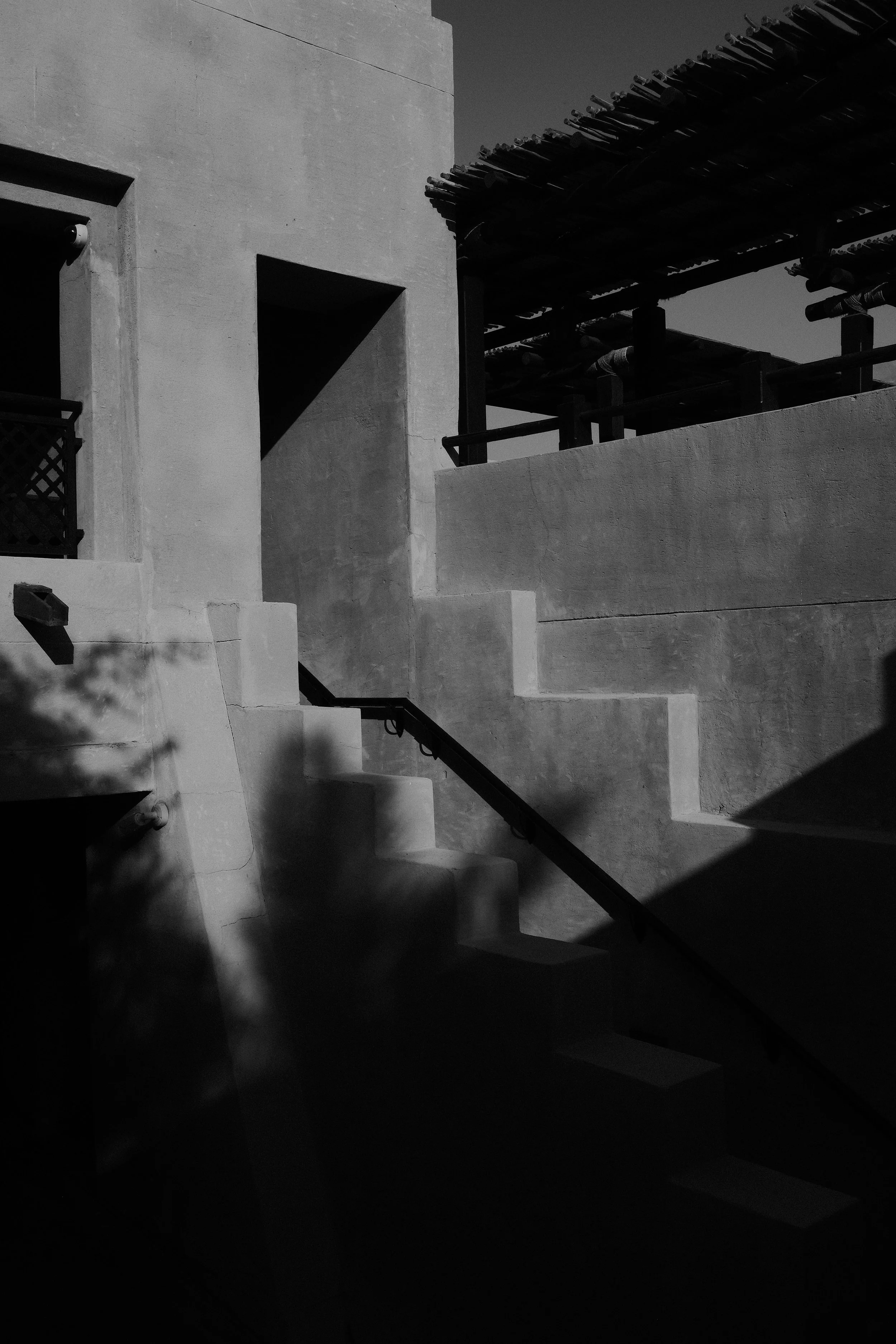 Black and white photo of a building's exterior with multiple staircases, walls, and a roof structure, casting shadows.
