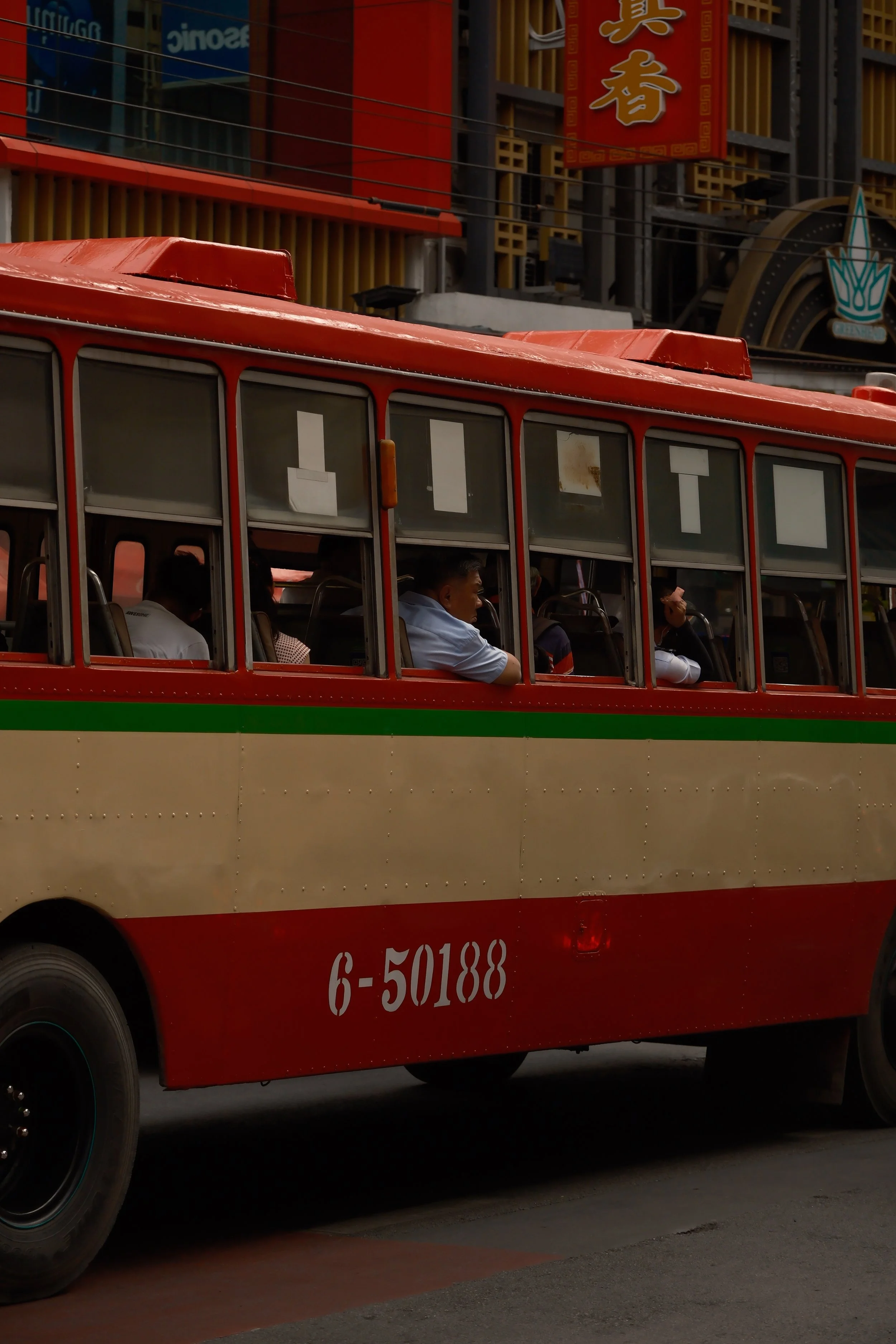 A red and cream bus with the number 6-50188 on the side, traveling on a city street. In the background, there are illuminated signs and traditional Asian architectural elements, including a sign with Chinese characters and a stylized lotus flower log