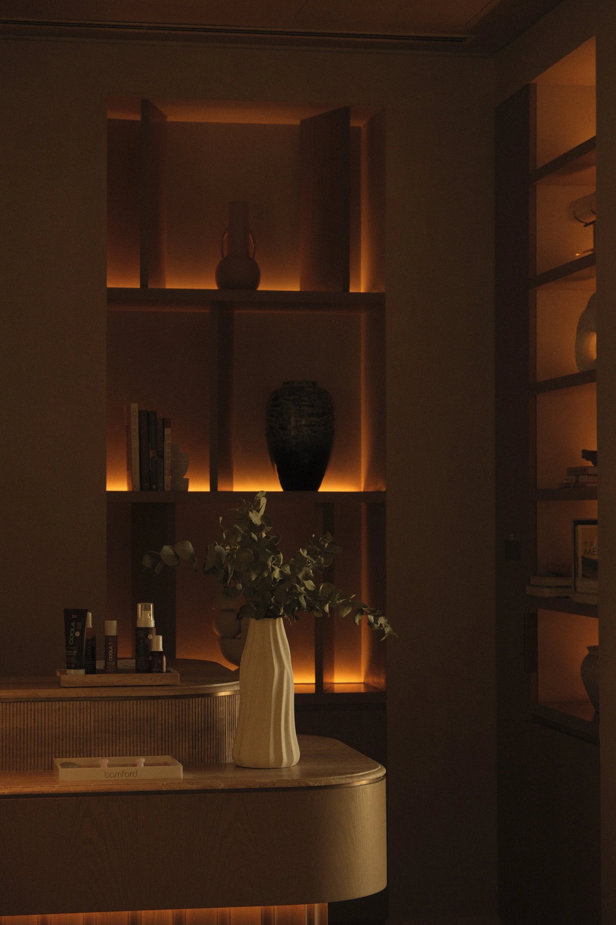 Dimly lit shelving unit with decorative vases and books, illuminated by warm backlighting, near a table with a white vase holding eucalyptus branches, in a cozy interior space.