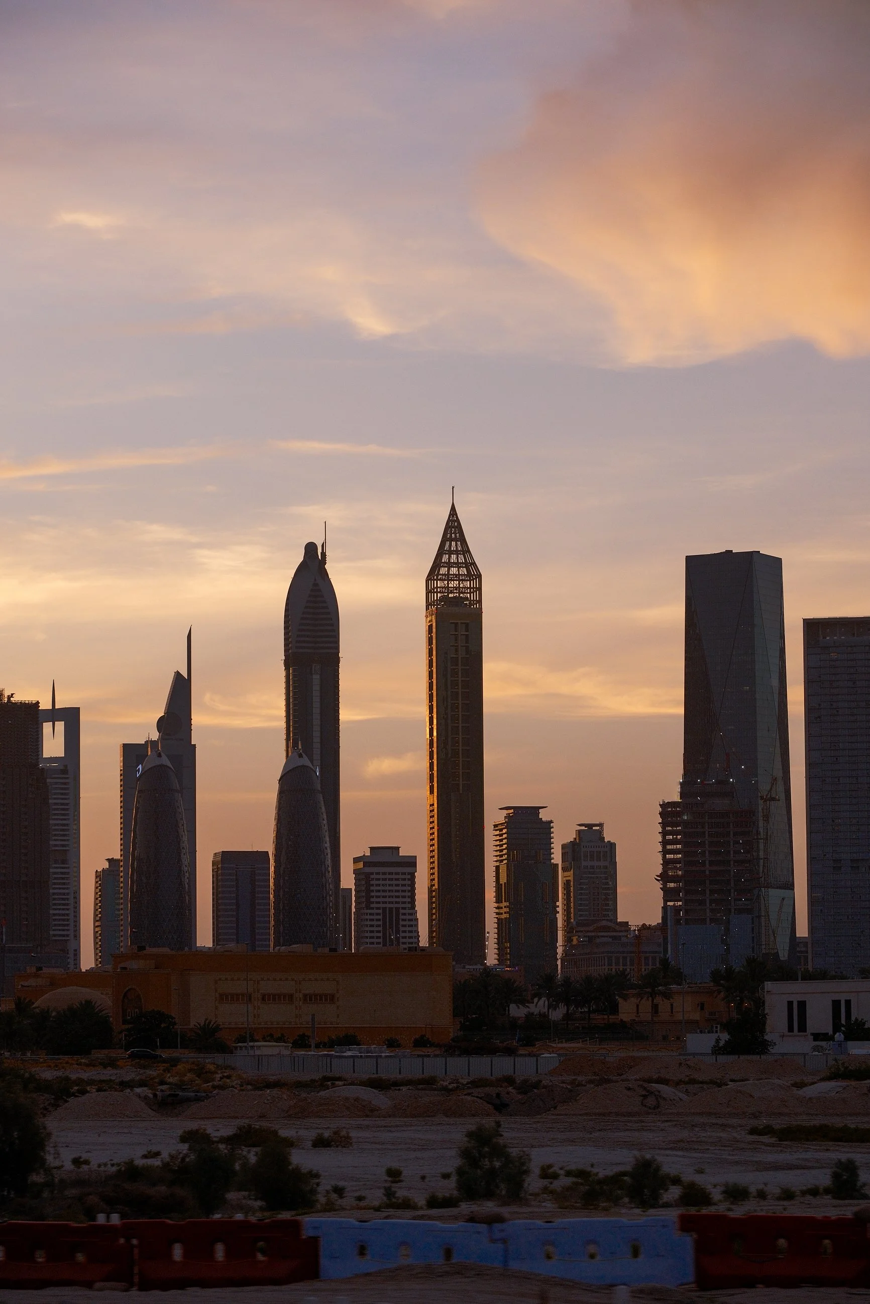 Sunset over a city skyline with modern skyscrapers, including two uniquely shaped towers, under a colorful sky with soft clouds.