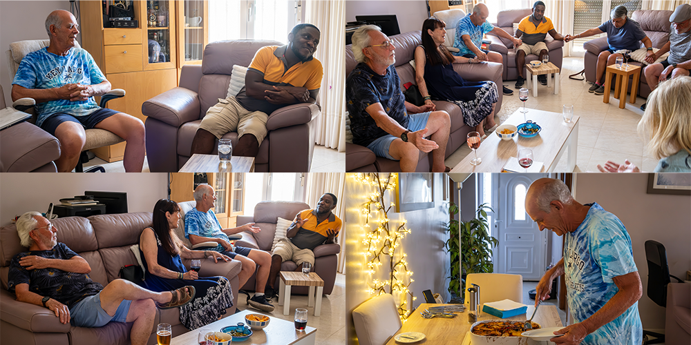 Group of people socializing in a living room with couches, drinks, and snacks, and a man with dinner in a dining area decorated with string lights.