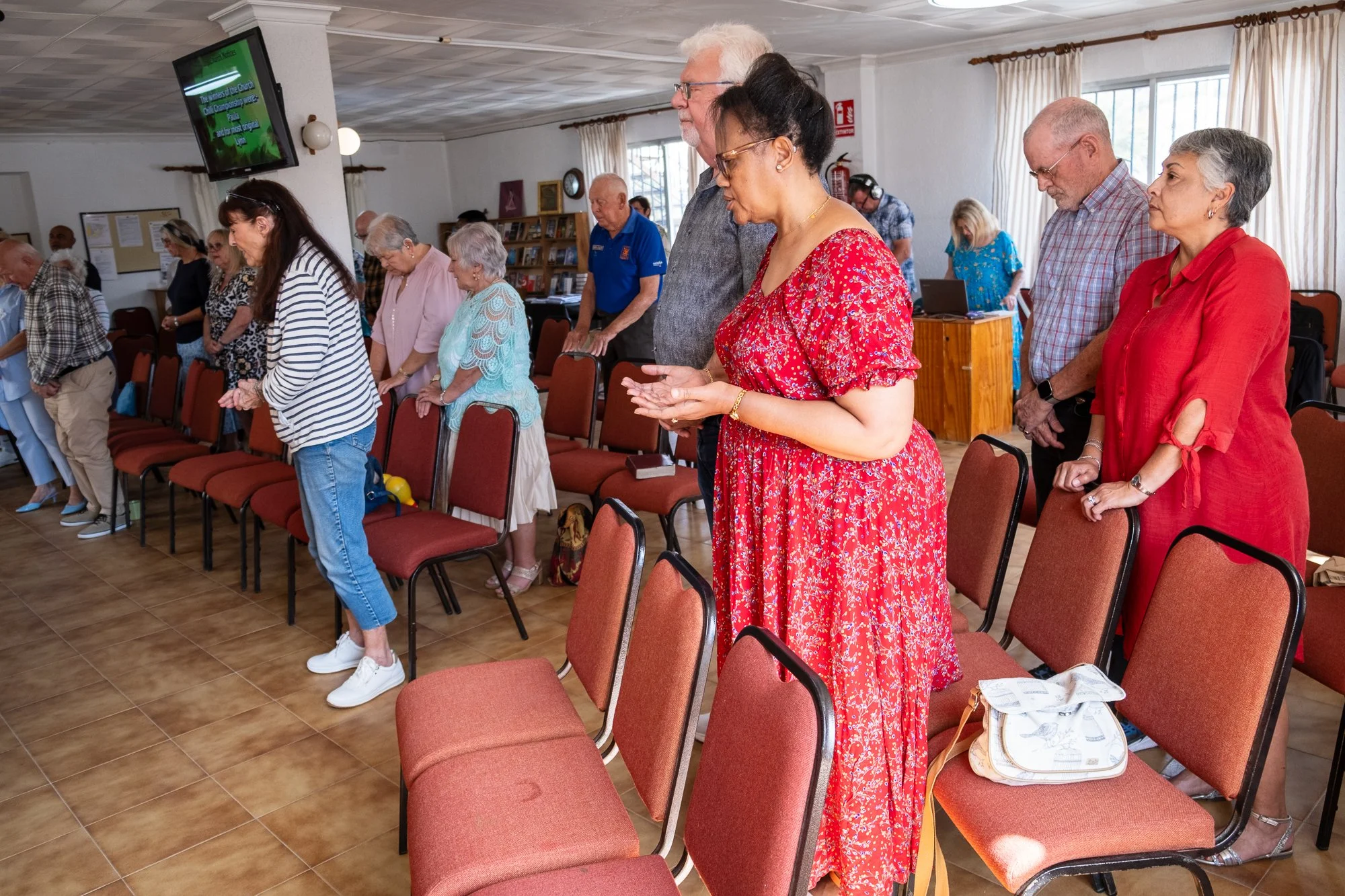 People are standing in a row with heads bowed in prayer or reflection in a church, and a television mounted on the wall.