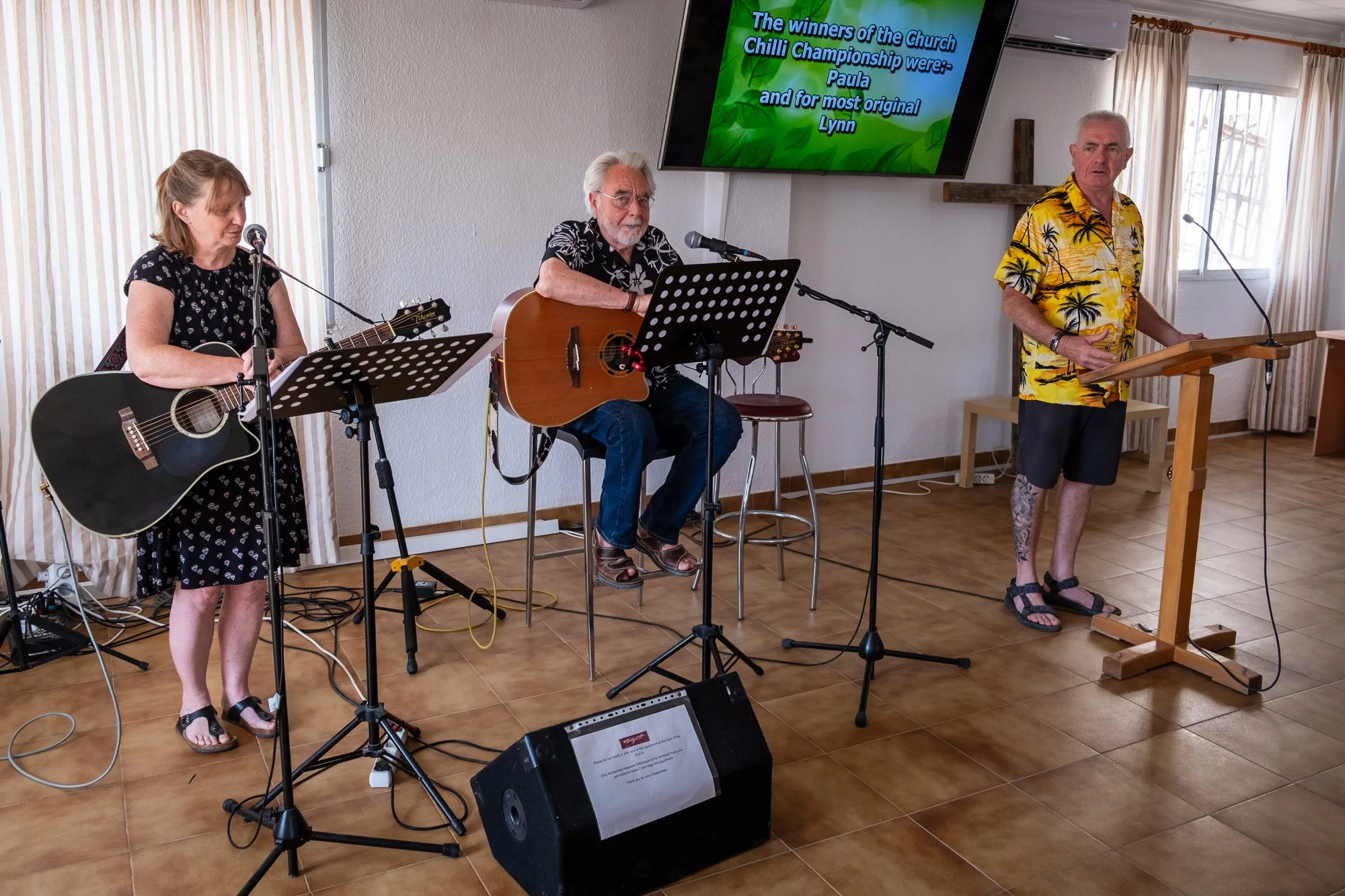 Three older adults performing music in a room with natural light—two women and one man. The woman on the left is playing an acoustic guitar, the man in the middle is also playing a guitar, and the man on the right is standing at a lectern, possibly singing or speaking. A large screen on the wall shows a message about church winners of a chili championship, and there is musical equipment, including microphones and speakers, on the floor.