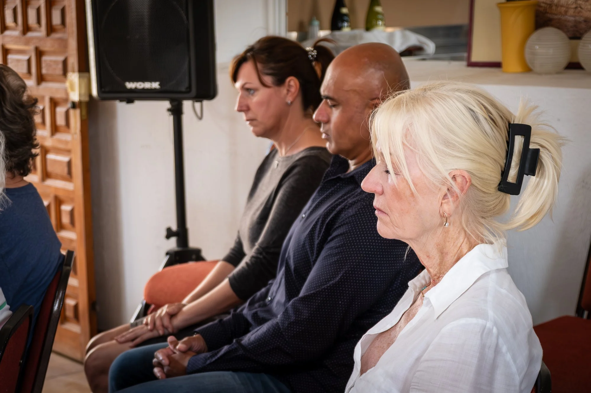 Four people sitting with eyes closed, participating in a meditation or relaxation session in a church with neutral decor.