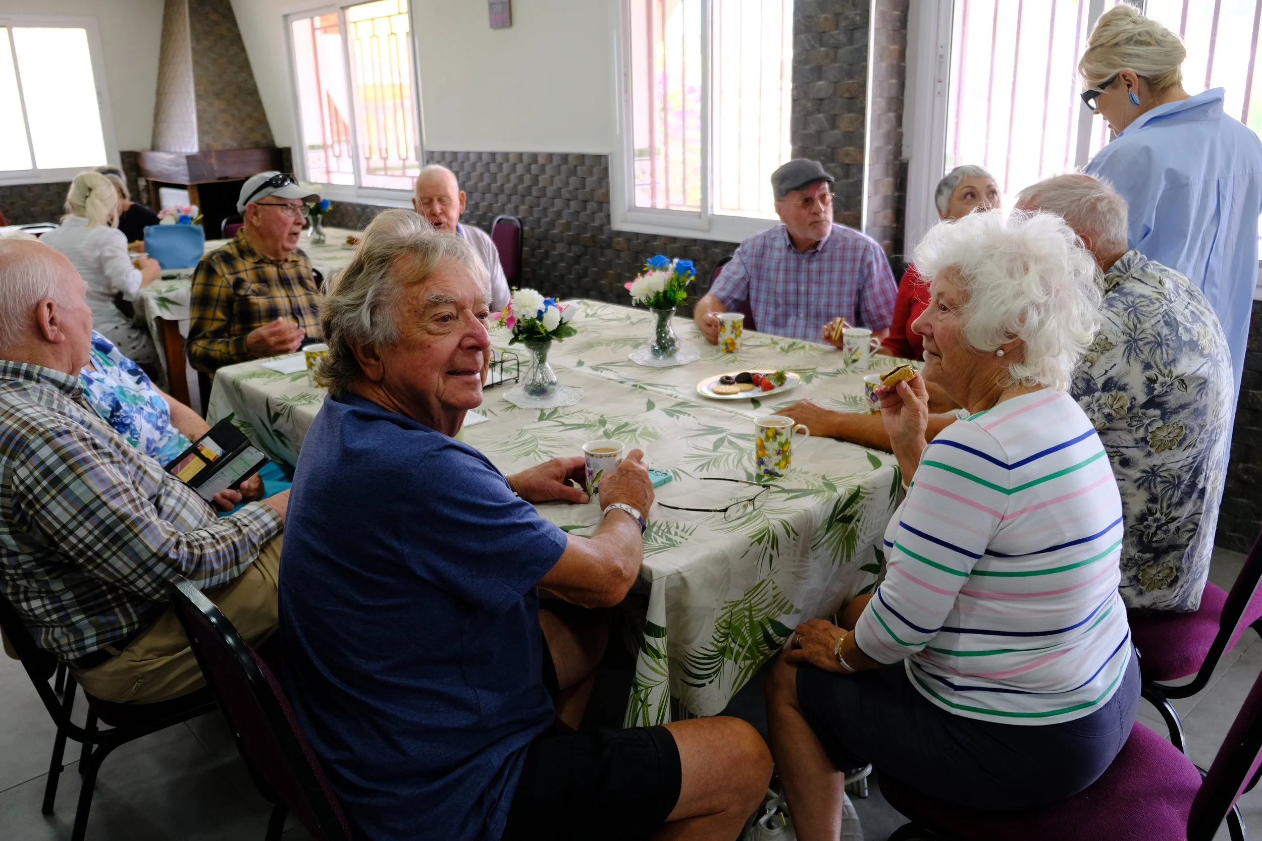 Group of elderly people sitting around a table with floral centerpiece, enjoying snacks and coffee in a bright room, while a woman standing on the right talks to them.