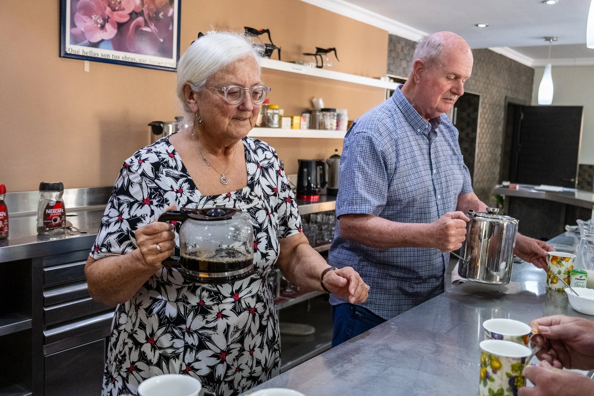 An elderly woman and man standing in a kitchen, pouring beverages into cups. The woman has white hair, glasses, and is wearing a black and white floral dress. The man has a bald head and is wearing a blue checkered shirt.