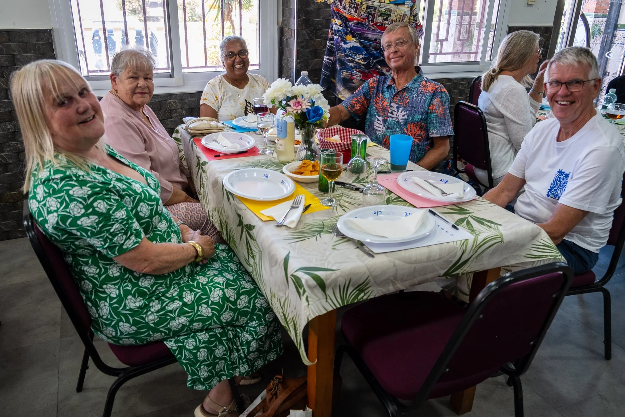 Group of six older adults sitting at a festive dinner table, smiling and enjoying a meal. The table is decorated with a floral centerpiece, plates, glasses, and snacks. The setting appears to be indoors with large windows in the background.