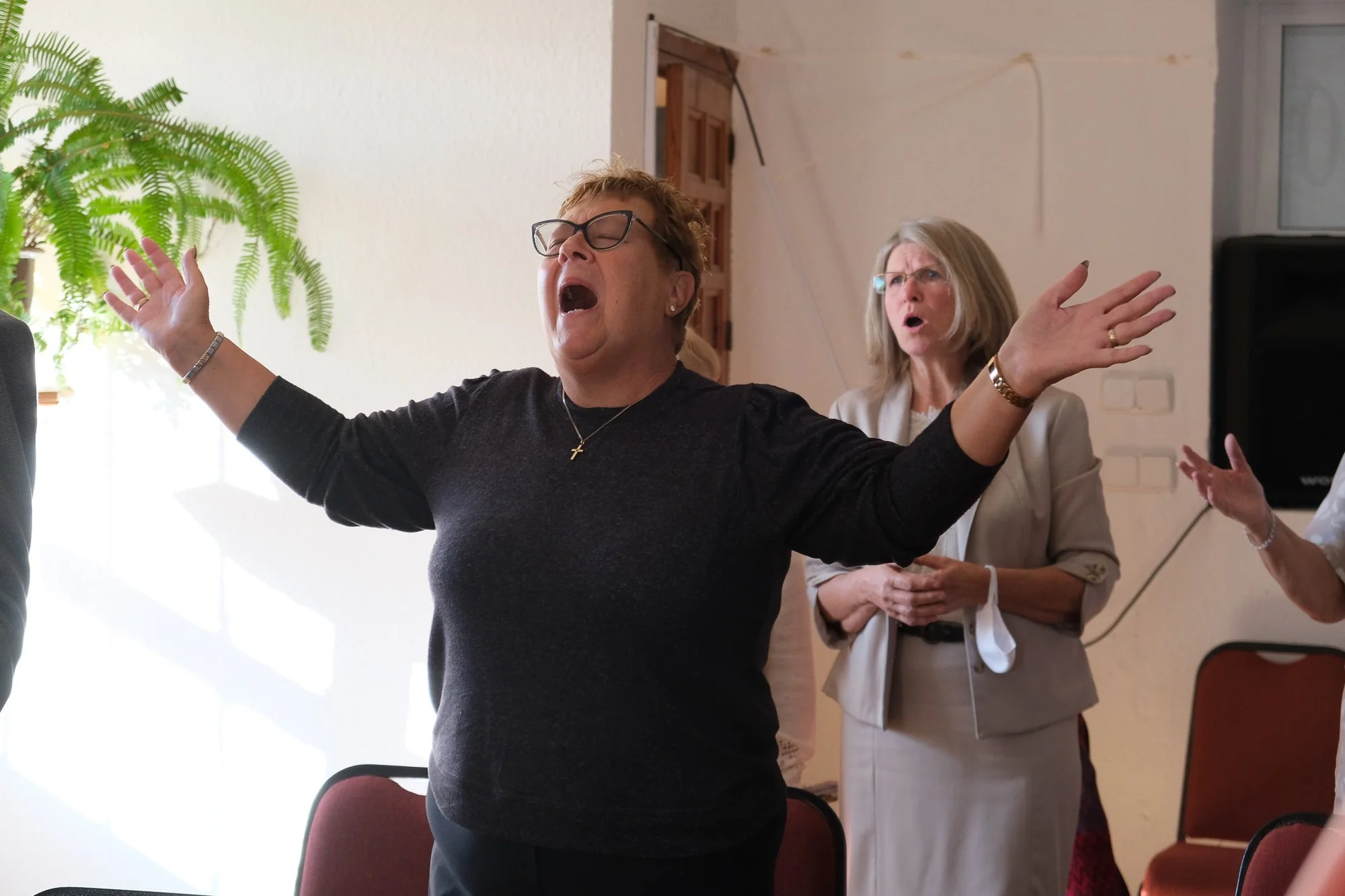 A woman with short curly hair, wearing glasses and a black shirt, appears to be singing or speaking passionately with her arms wide open. She is wearing a necklace with a cross pendant. Another woman with shoulder-length gray hair and glasses, dressed in a light-colored blazer, stands behind her and appears to be reacting emotionally.