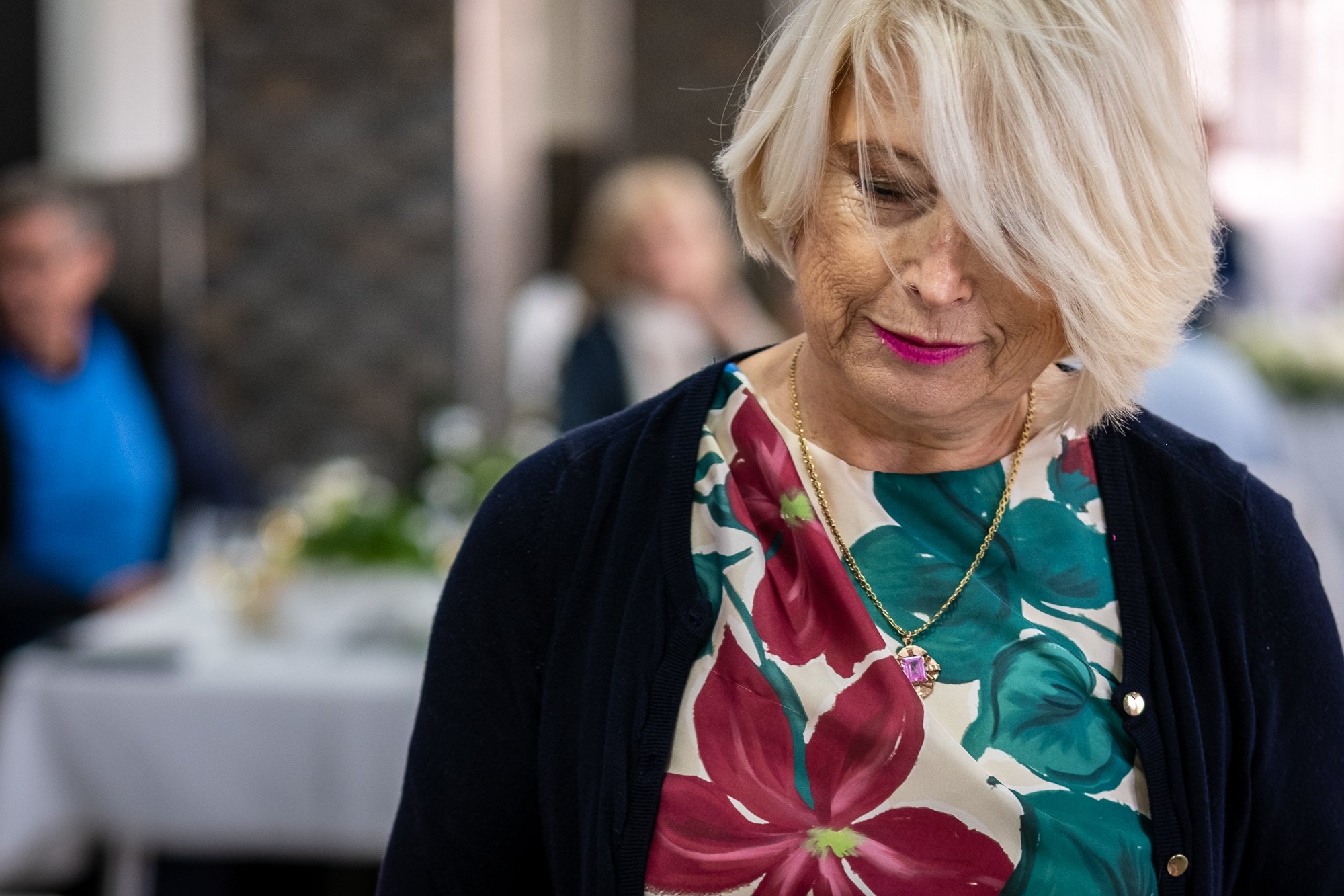 A woman with short platinum blonde hair, wearing a colorful floral dress and a black jacket, looking down and smiling gently.