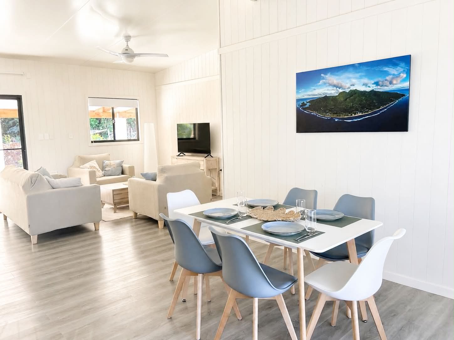 Open-concept living and dining area with white walls, a white ceiling fan, light wood flooring, a dining table set for six, and a cozy living room with white sofas facing a TV, with windows and sliding door letting in natural light.