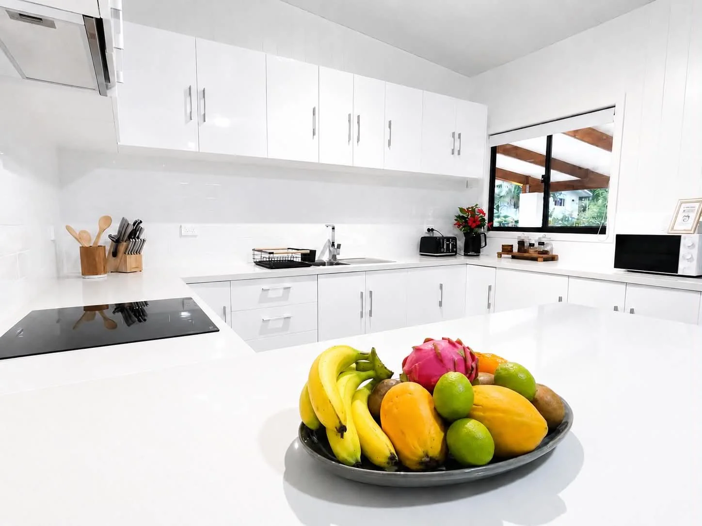 White kitchen with a bowl of assorted fruits including bananas, mangoes, limes, and dragon fruit on a white countertop, and a window showing trees outside.