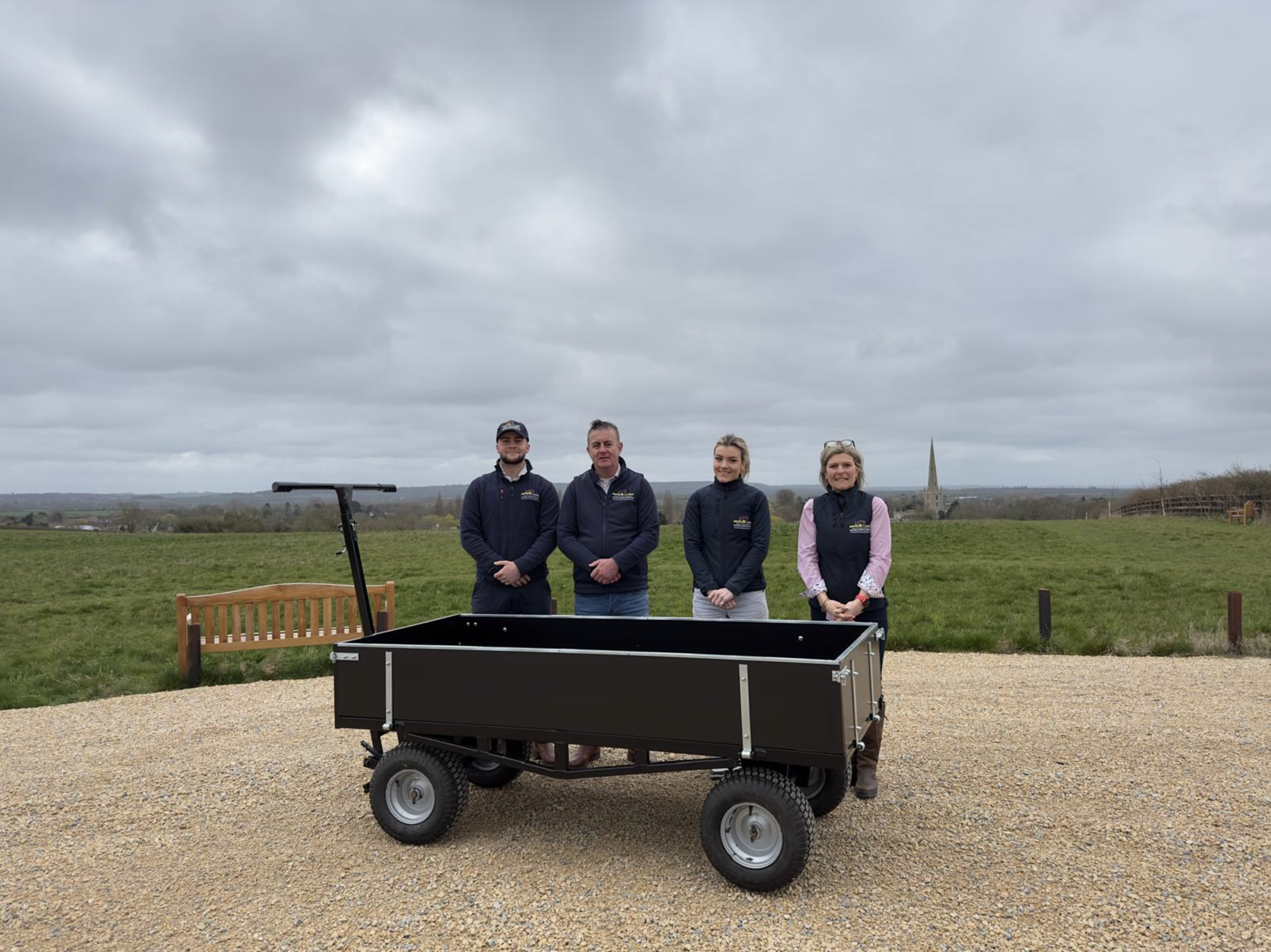 Four people standing outdoors on a cloudy day behind a funeral bier cart with large wheels, with a scenic landscape and church steeple in the background.