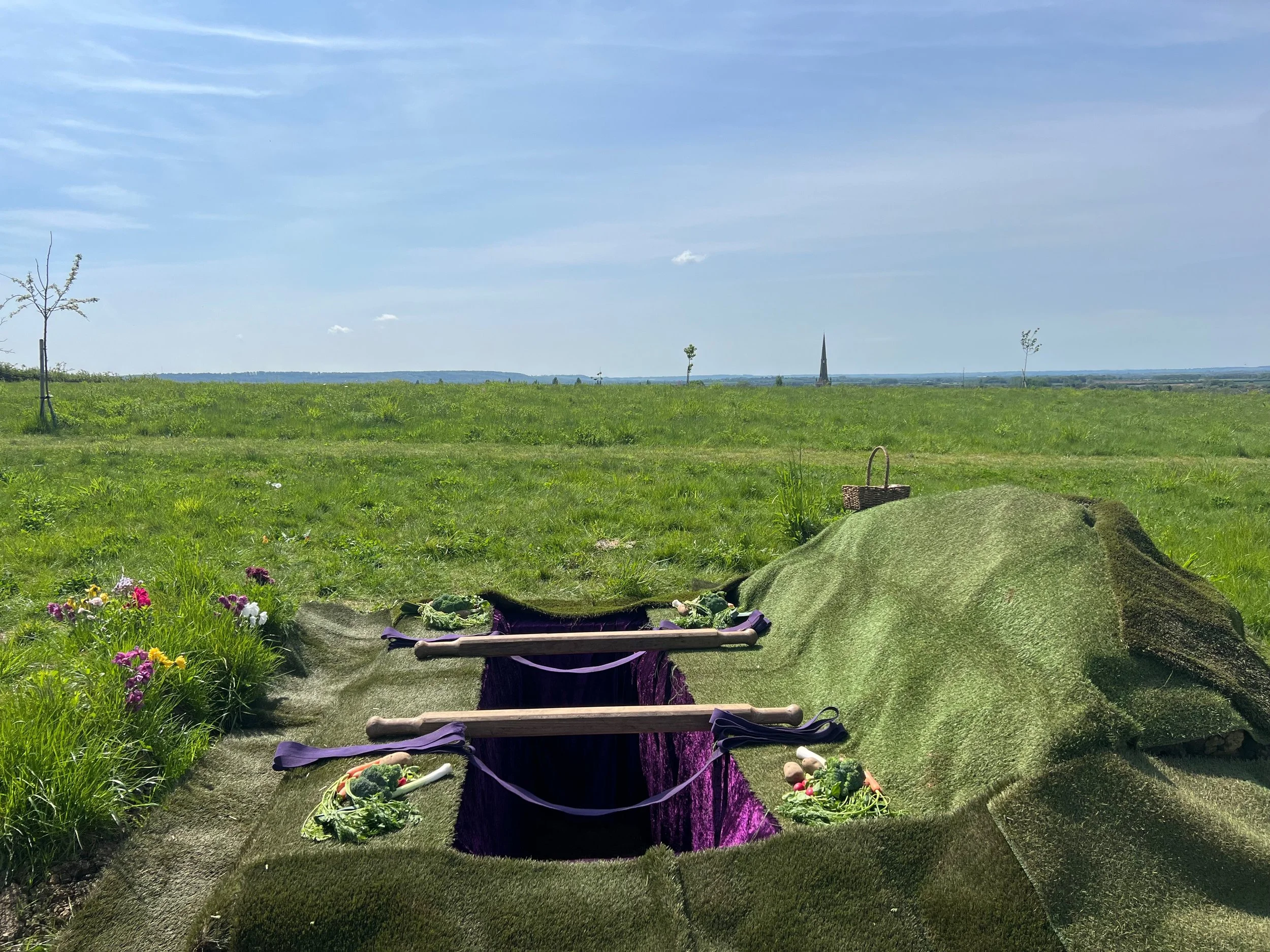 A grassy field with a few trees under a blue sky, with a distant church steeple on the horizon and a small garden bed in the foreground. There is a woven basket on the grass and a green mat in the foreground with some wrapped objects and purple fabric.