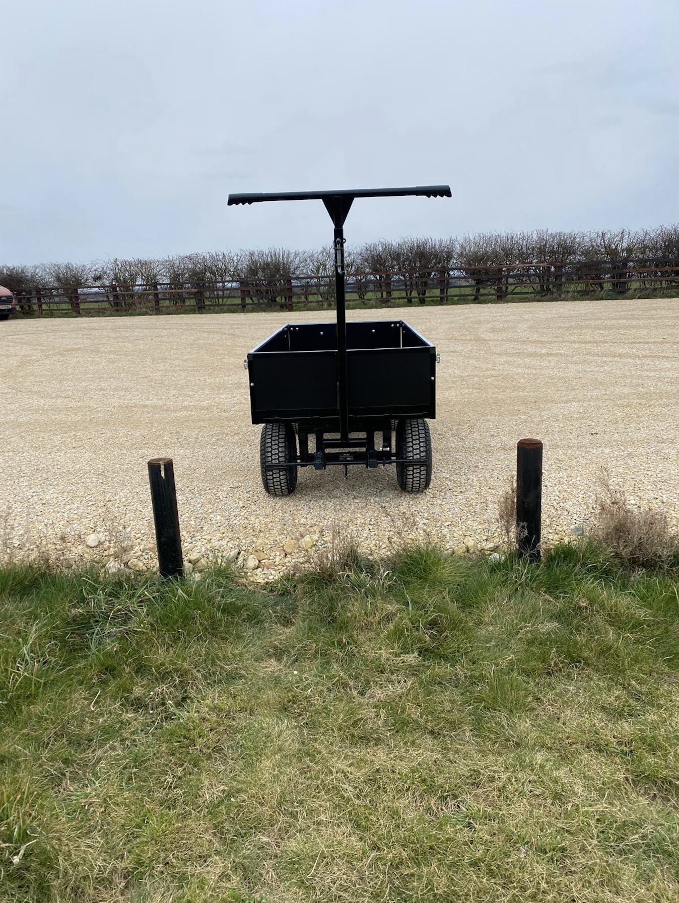 Black funeral bier cart with a raised handlebar on a gravel surface, with a grassy area and a wooden fence in the background.