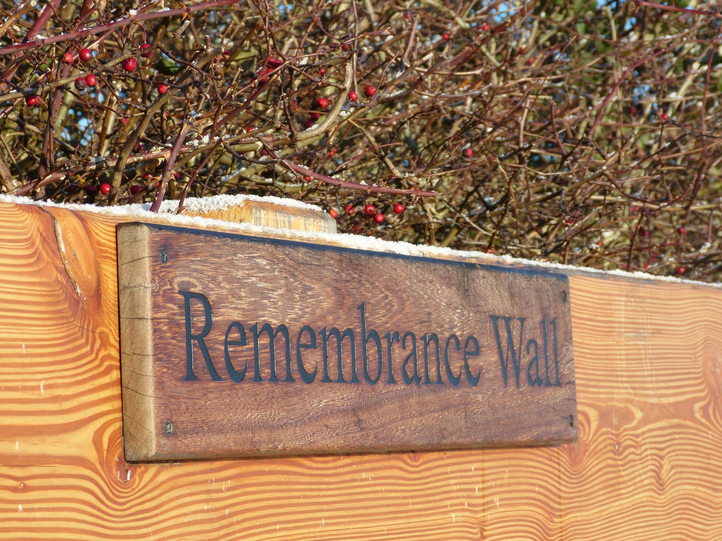 Close-up of a wooden sign with the words 'Remembrance Wall' engraved on it. The sign is mounted on a wooden surface, with some snow on top. In the background, there are dense, leafless branches with small red berries, and a bright sky visible through the branches.