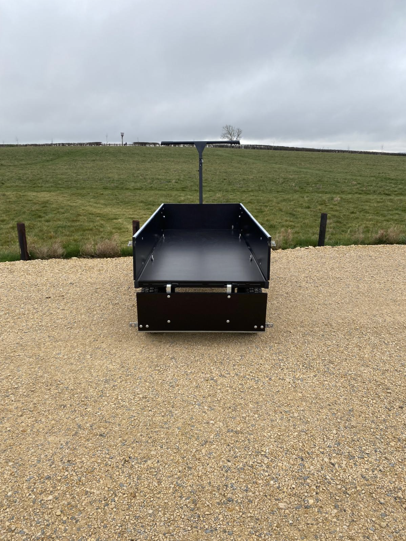 A black metal funeral bier cart on a gravel carpark in front of a grassy field under a cloudy sky.