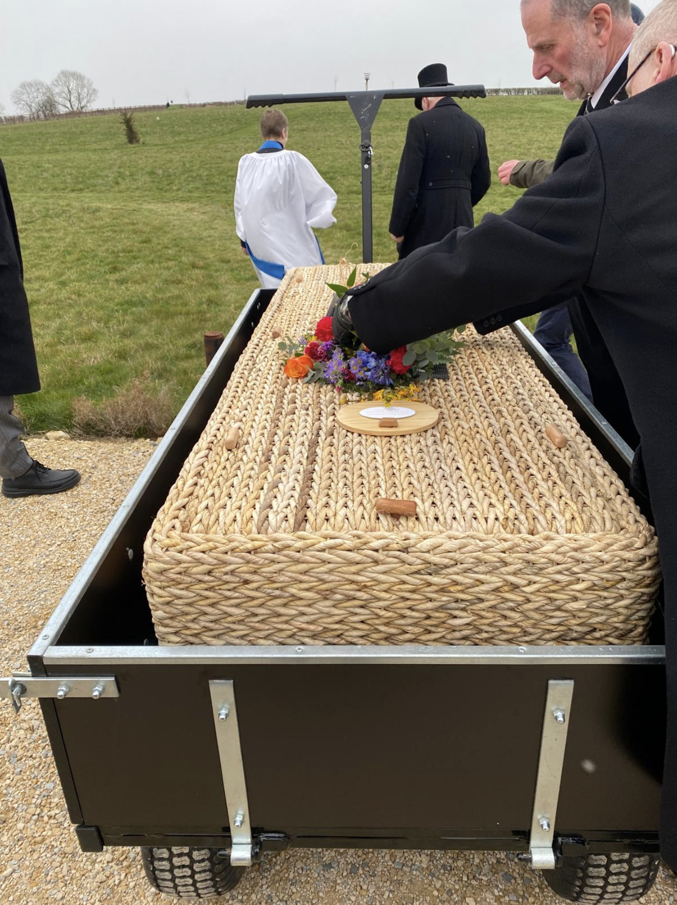 A coffin covered with a woven basket-like material on a metal cart, surrounded by people, some in formal clothing, in an outdoor setting with grassy fields and a cloudy sky.