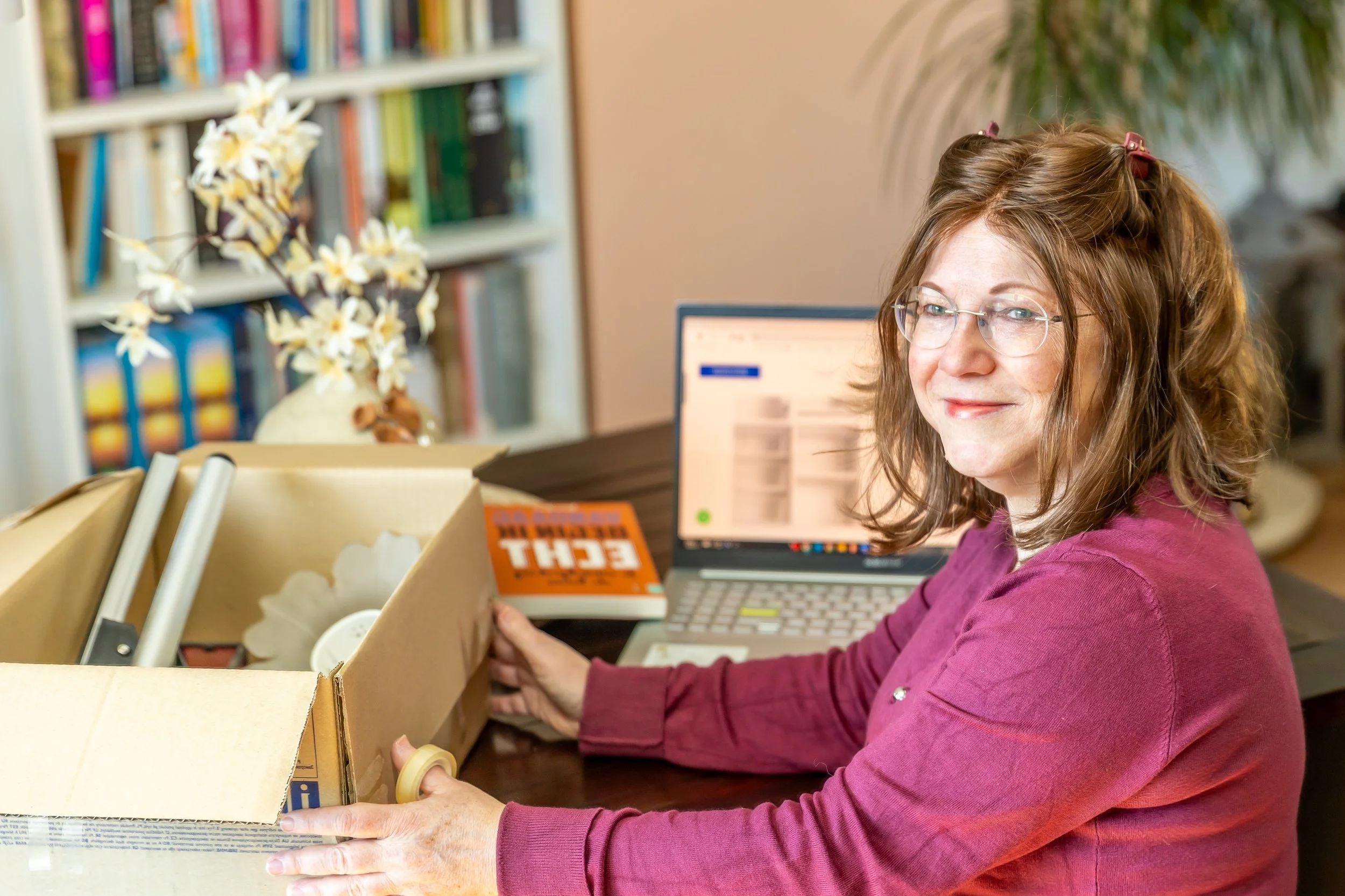 Een vrouw met rood haar en bril die in een kamer zit, omringd door boeken en een computer, terwijl ze een doos met vinylplaten vasthoudt.