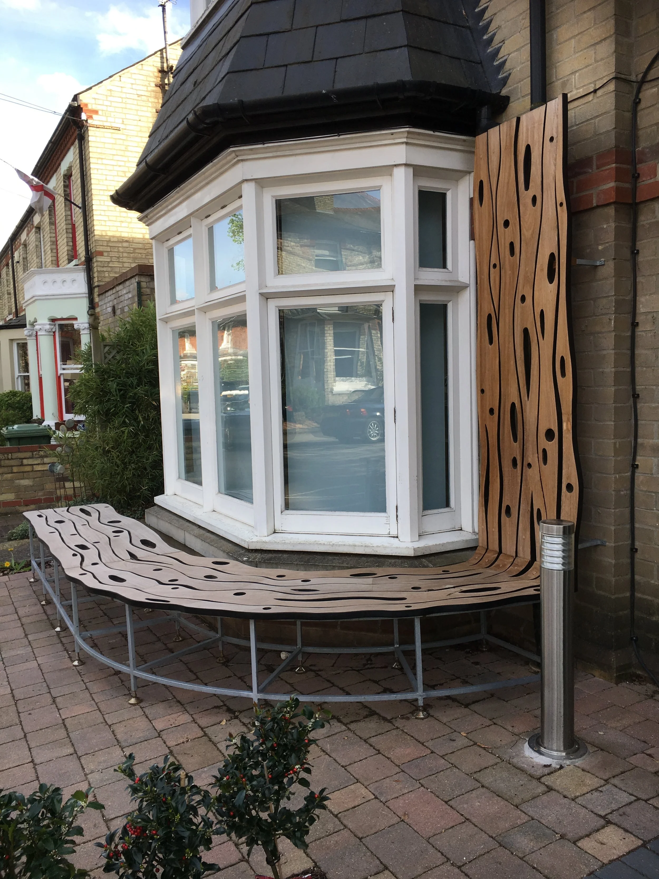 A decorative architectural feature with a large, curved wooden bench with a unique organic cutout pattern, situated outside in front of a house with brick walls and a bay window. A modern metal pole with a built-in heater or light is next to the bench, and there are small bushes and a brick paved pathway in the foreground.