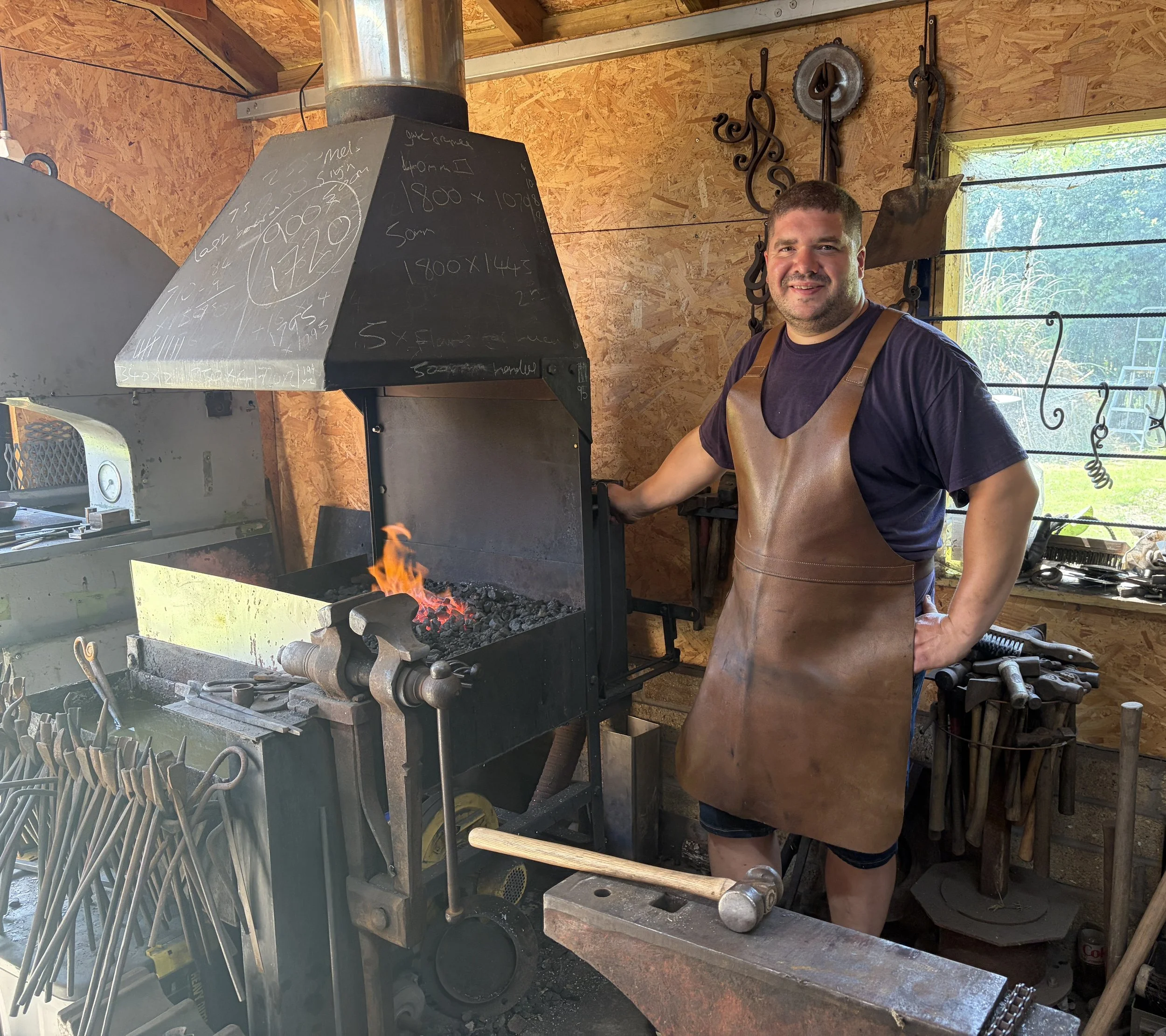 A smiling man wearing a leather apron working on an anvil in a blacksmith workshop with tools hanging on the wooden wall behind him and a large window showing greenery outside.