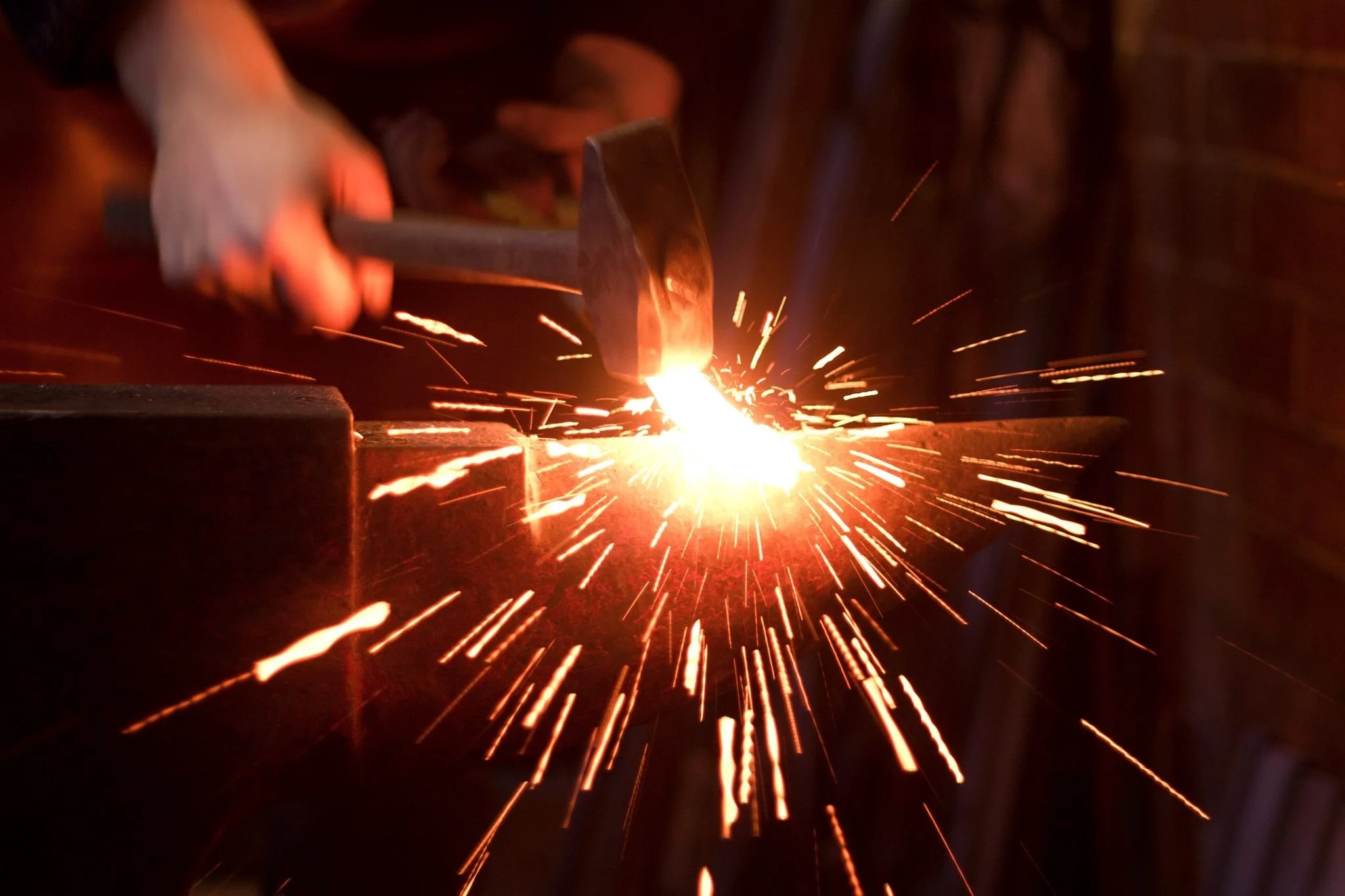 A hand striking metal with a hammer near a spark-producing welding or forging process in a workshop.