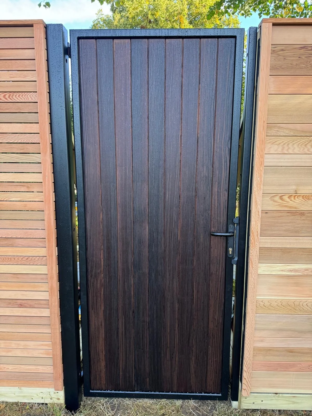 A wooden gate with vertical panels, black frame, and handle, installed between two wooden fences.