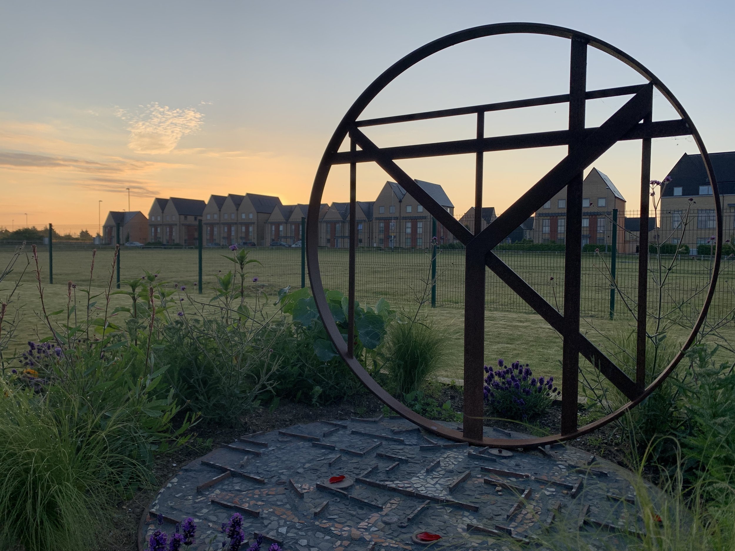 A decorative metal sculpture of a bicycle wheel in a garden at sunset, with flowers and a row of houses in the background.