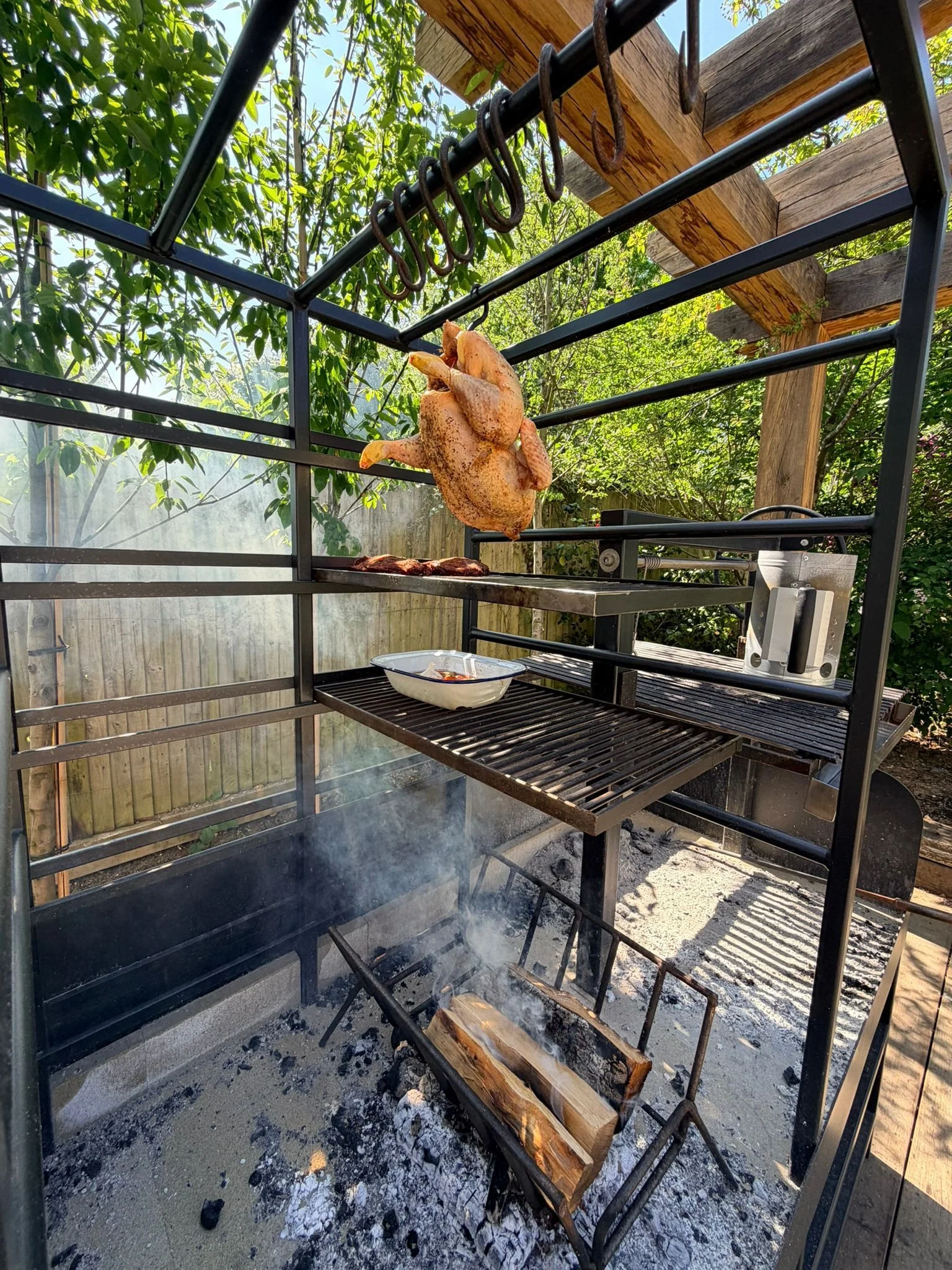 A whole roasted chicken hanging inside an outdoor meat smoker or grill, with a bowl below it, set against a background of trees and a wooden outdoor structure.