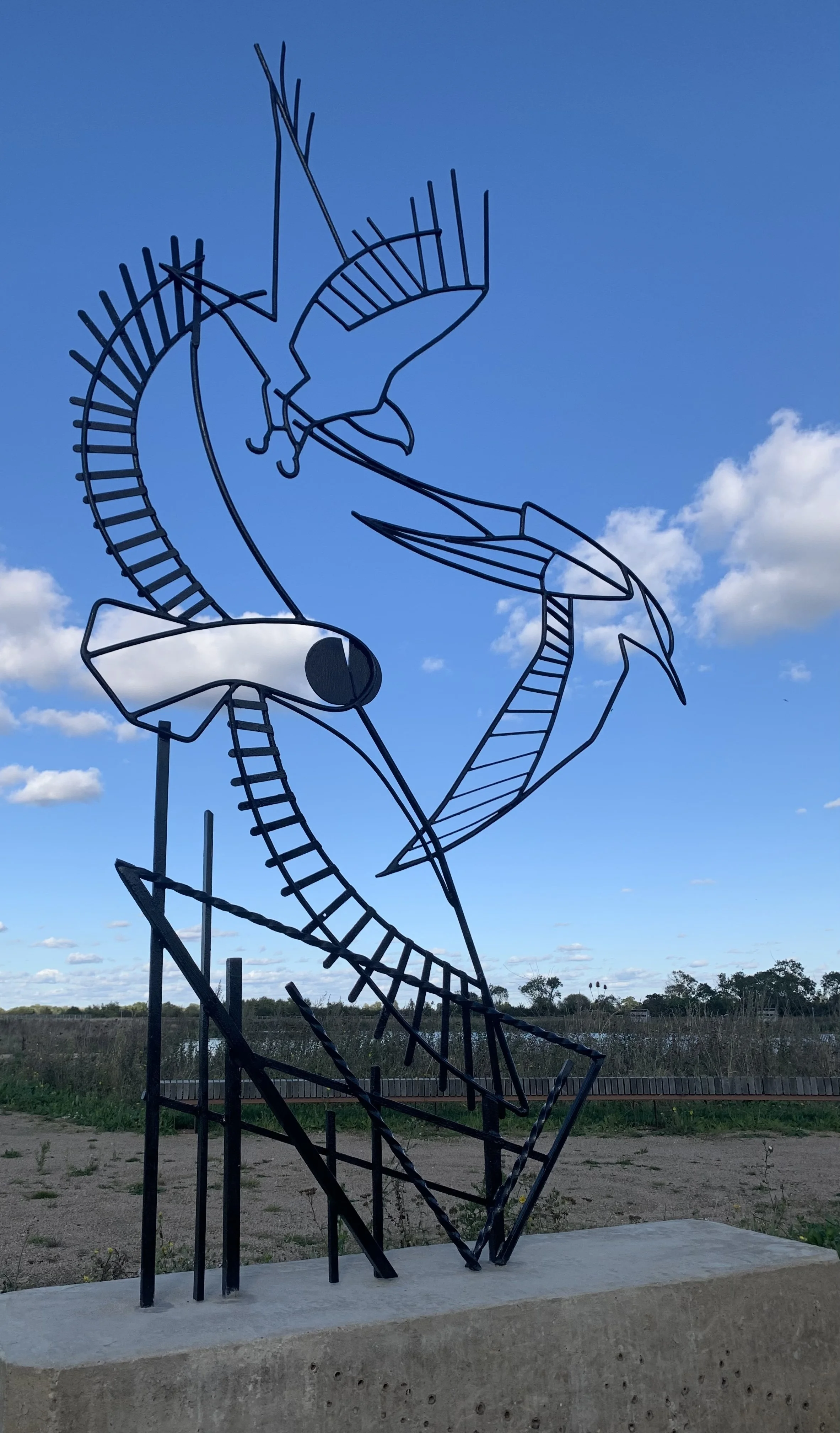 A metal sculpture of a stylized bird, possibly a heron or egret, with elongated neck and beak, set against a blue sky with scattered clouds, located outdoors on a concrete base.