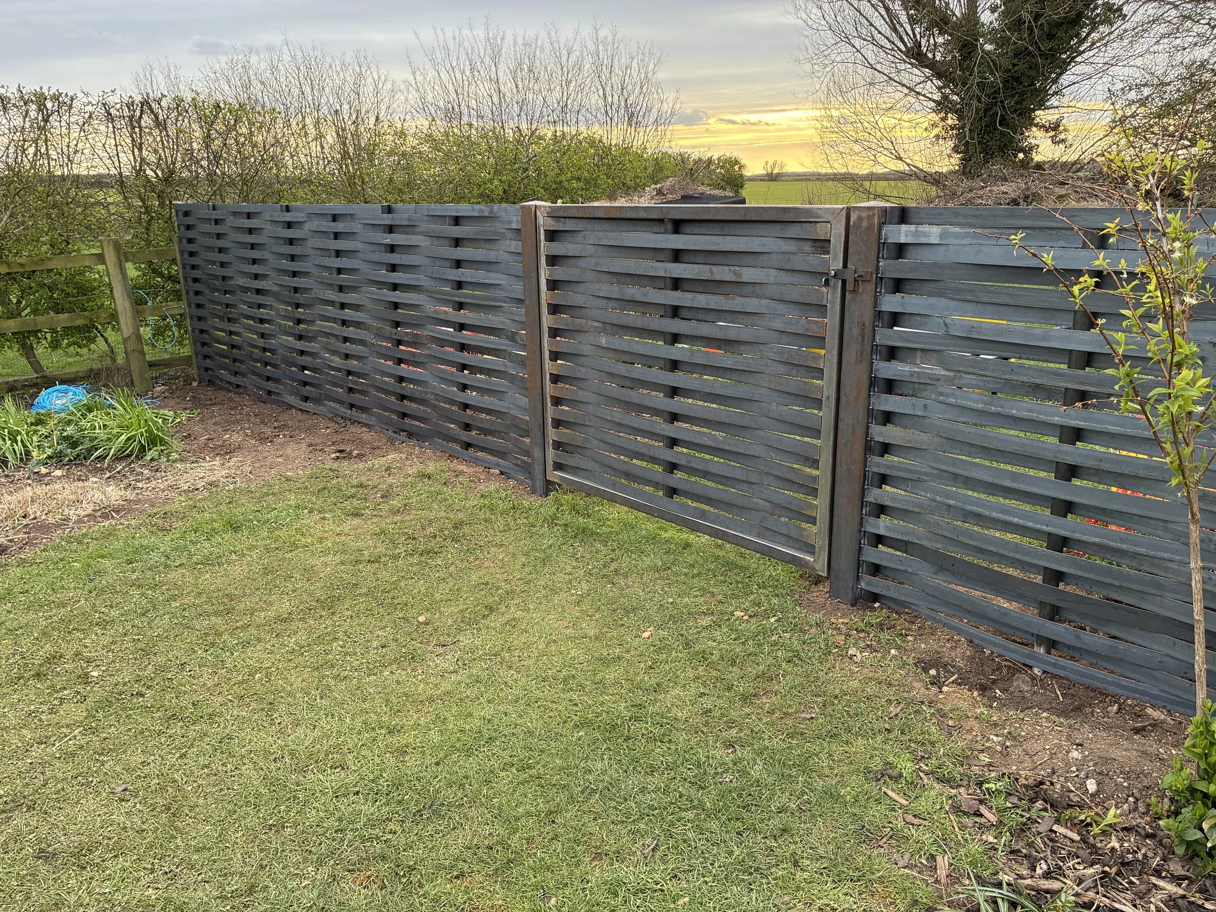 A black wooden fence with a latch in a field with green grass, trees, and a sunset sky in the background.