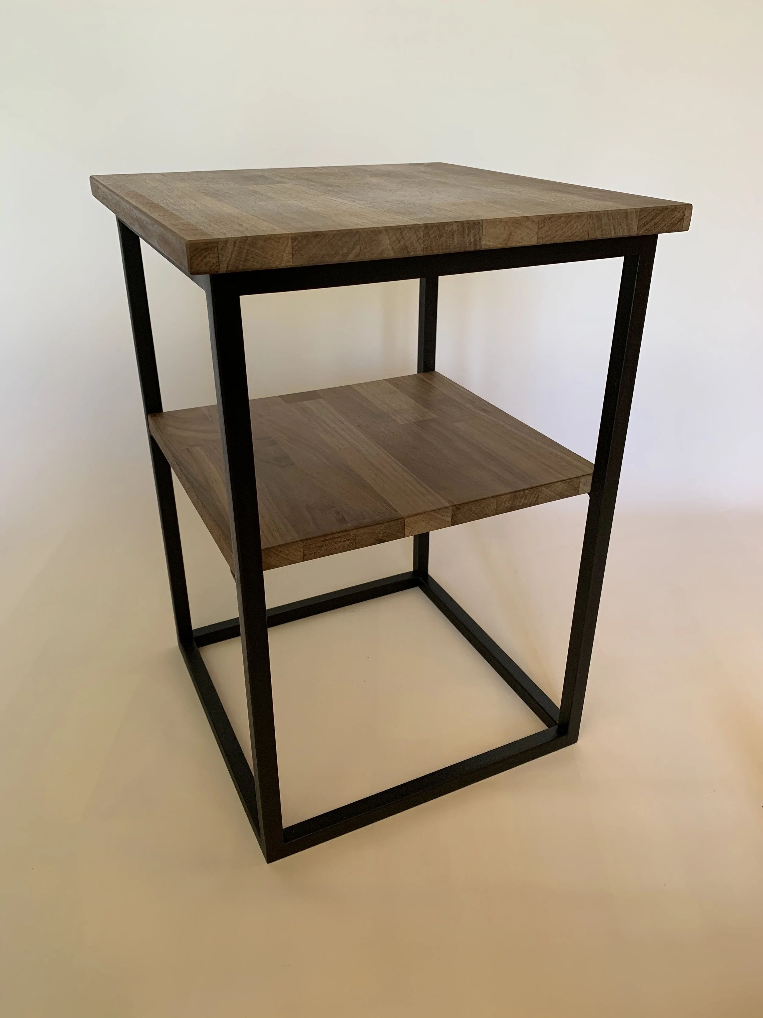 A small, three-tiered side table with a black metal frame, a wooden top, and a wooden lower shelf, placed against a plain light-colored background.