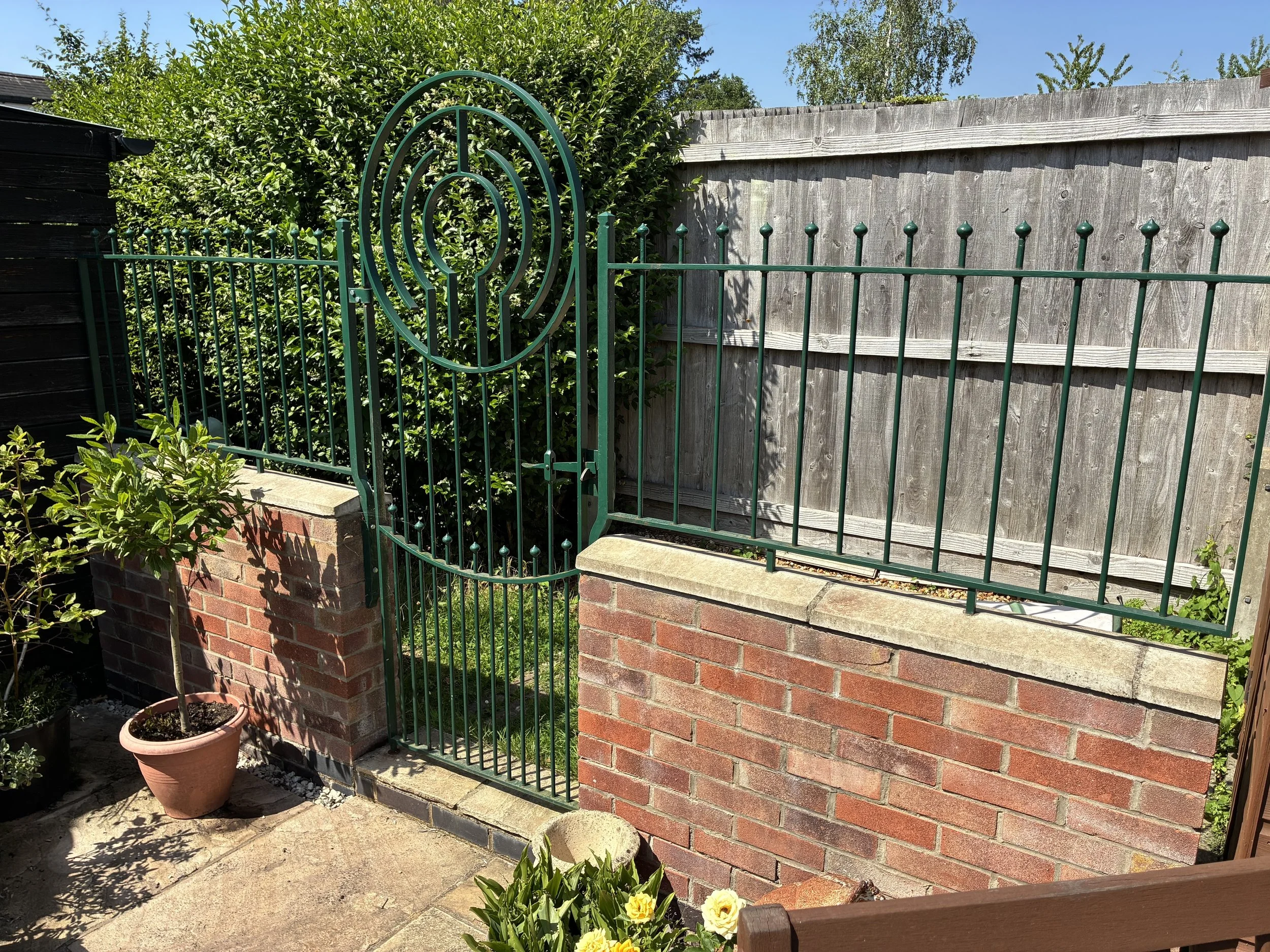 A brick fence with iron gate and green bushes in a backyard garden.