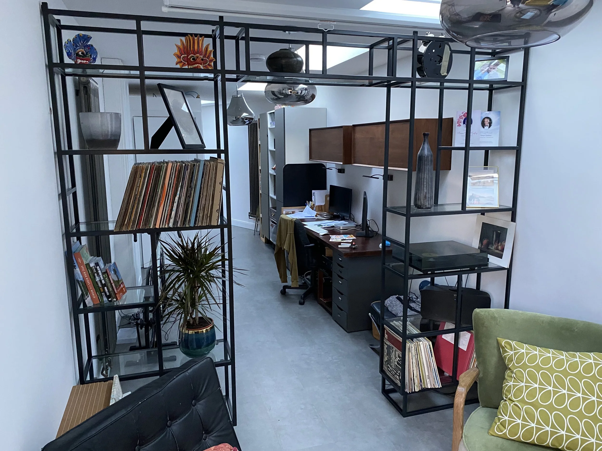 Living room with black metal open shelving containing decorative masks and vases, a plant, and vinyl records, with a desk and computer in the background.