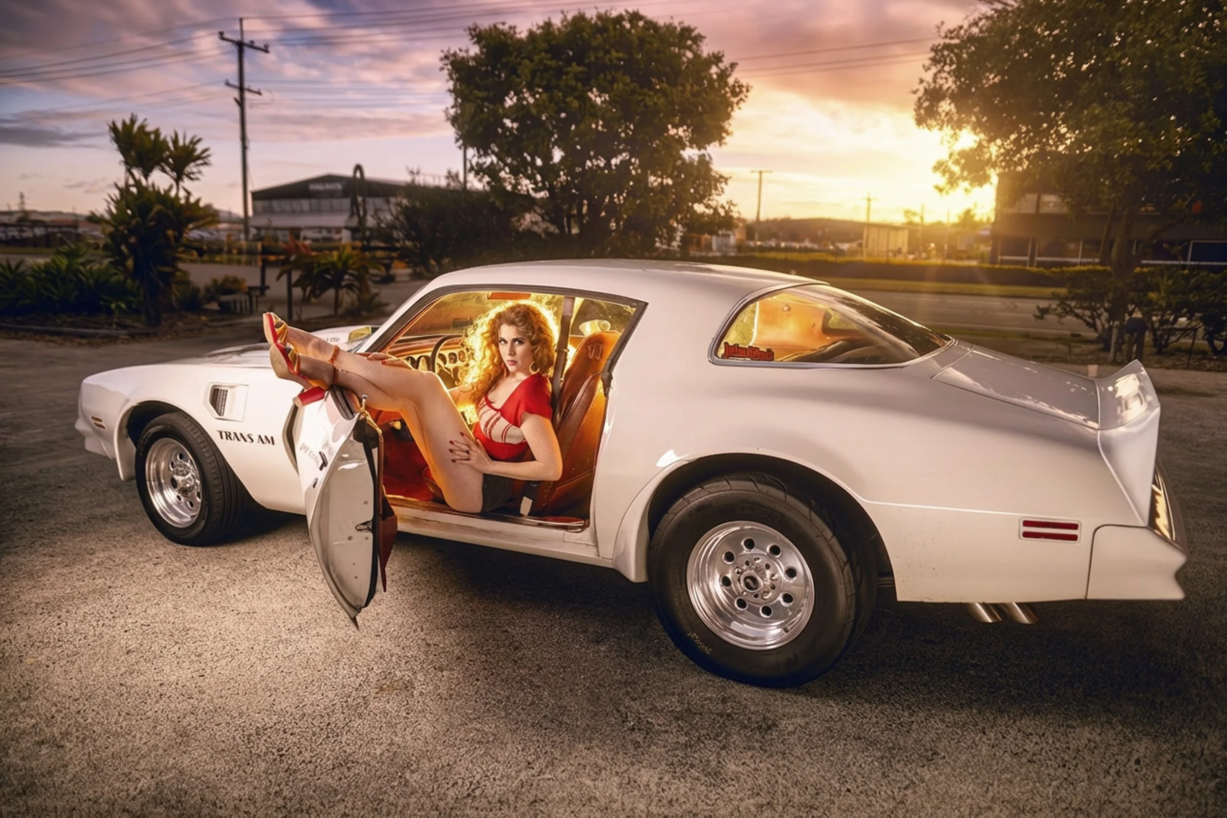A woman with curly red hair and a red dress sitting in a vintage white muscle car, with the driver's side door open, during sunset in a parking lot surrounded by trees and buildings.