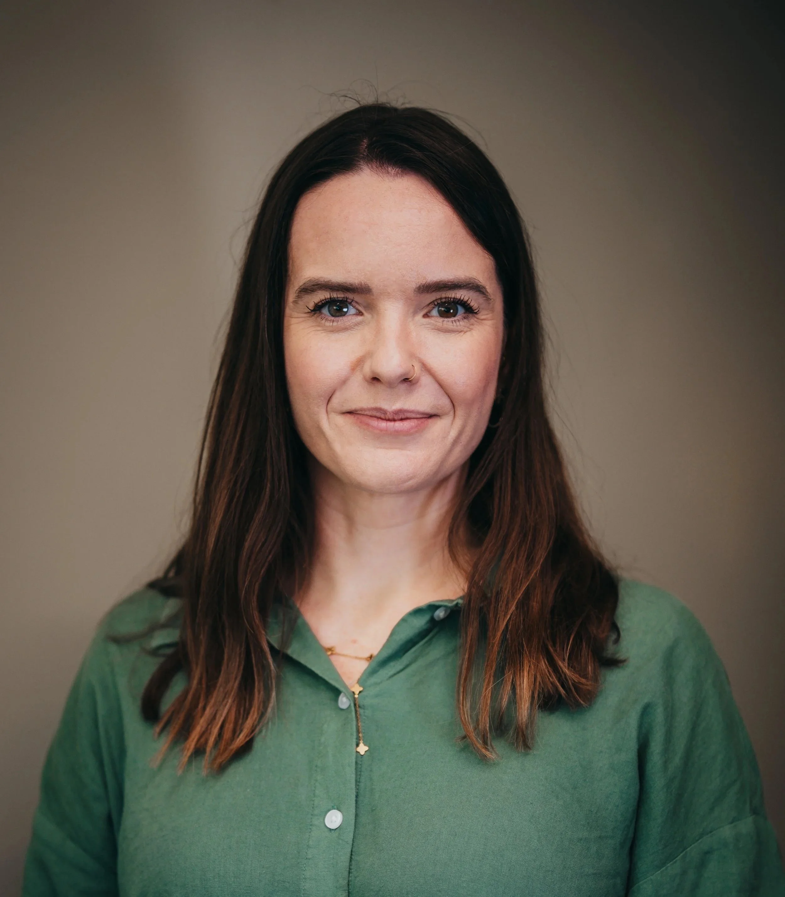 A woman with brown hair wearing a green sweater and a delicate gold necklace with a small cross pendant, smiling gently, with a neutral background.