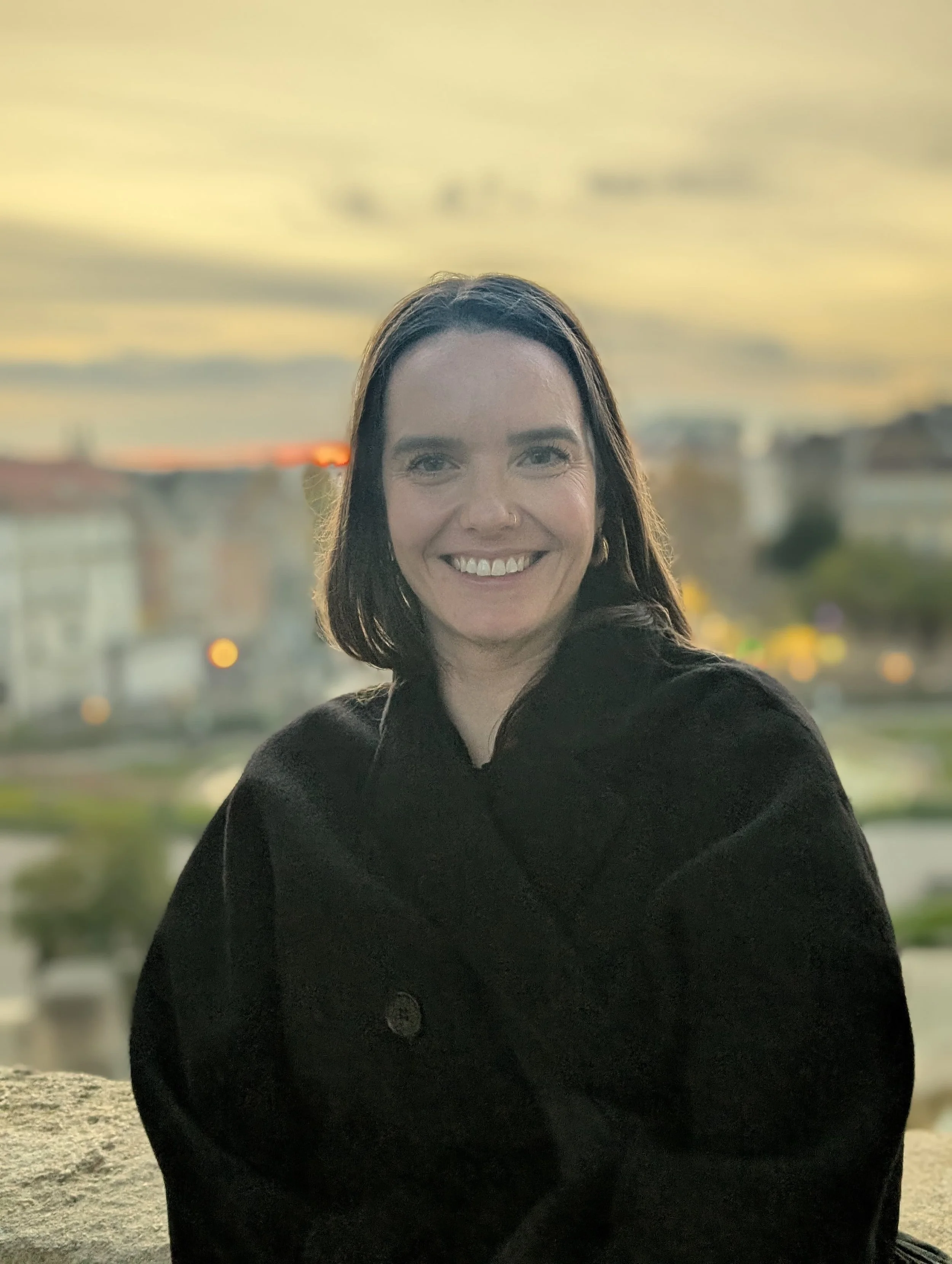 A woman with long dark hair, outdoors during sunset with a blurred cityscape background.