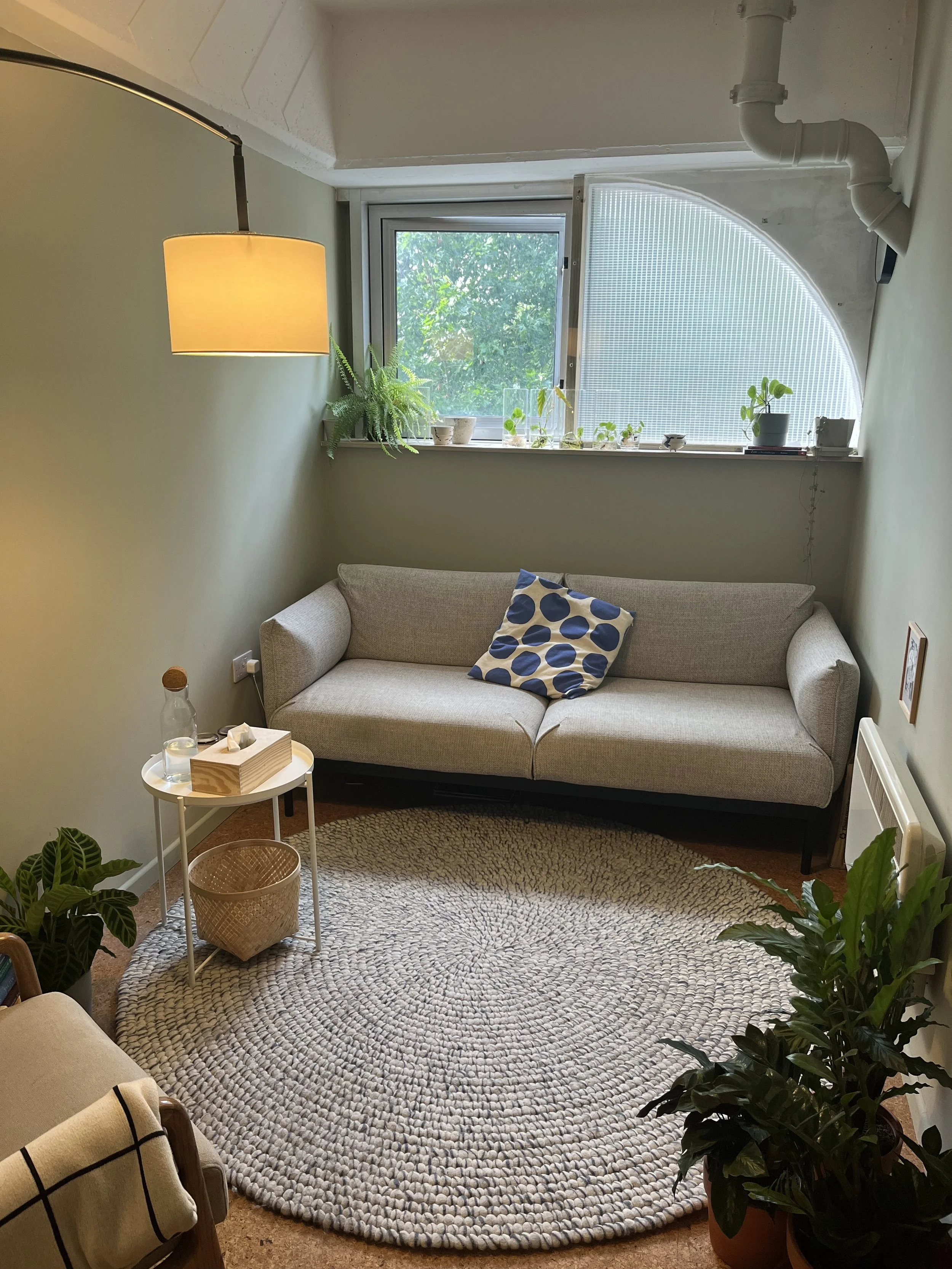 Living room with a beige sofa, potted plants on the window sill, a round woven rug, a side table with a tissue box and a glass, a yellow lamp, and a window with a semi-circular design and view of green trees outside.