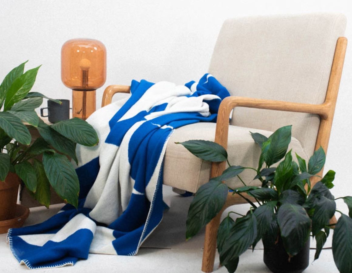 A beige armchair with wooden armrests and legs, draped with a blue and white striped blanket. There are potted plants on either side of the chair, a black mug on a side table, and an amber-colored table lamp with a glass shade.