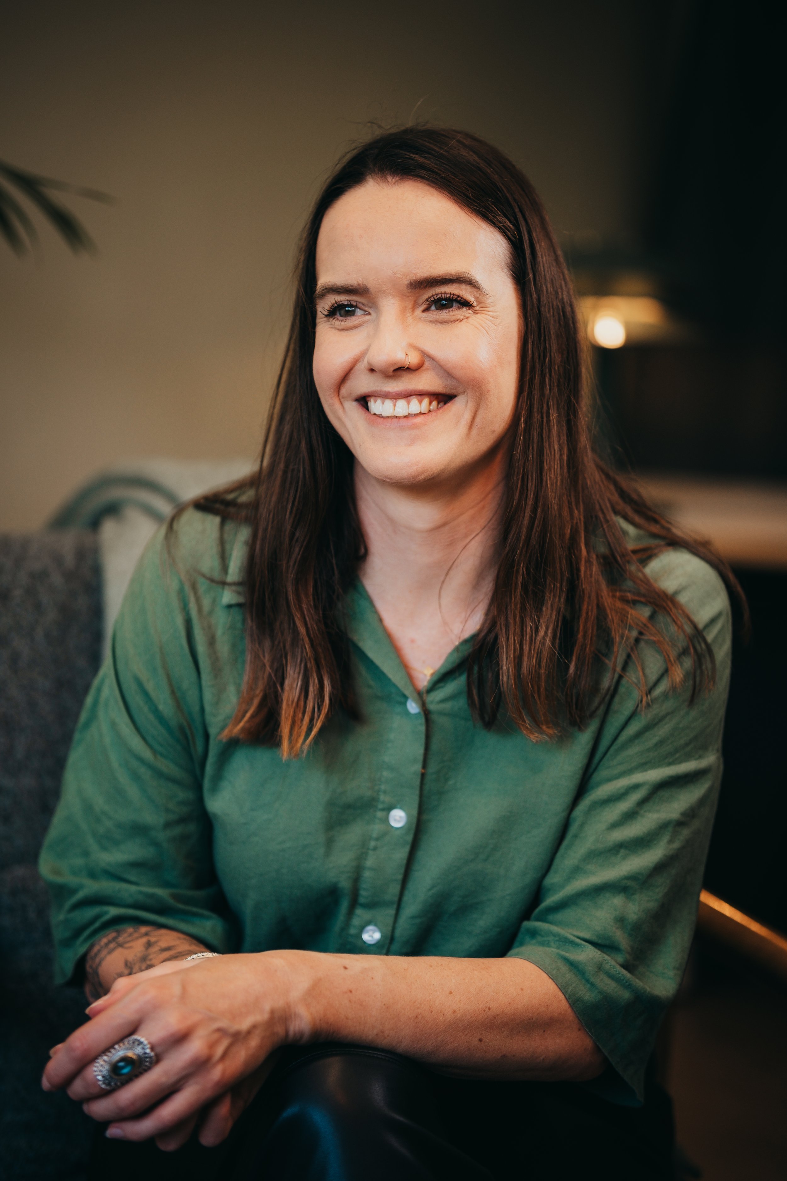 Close-up of a smiling woman with long brown hair wearing a green shirt, seated with her arms crossed on a dark table.