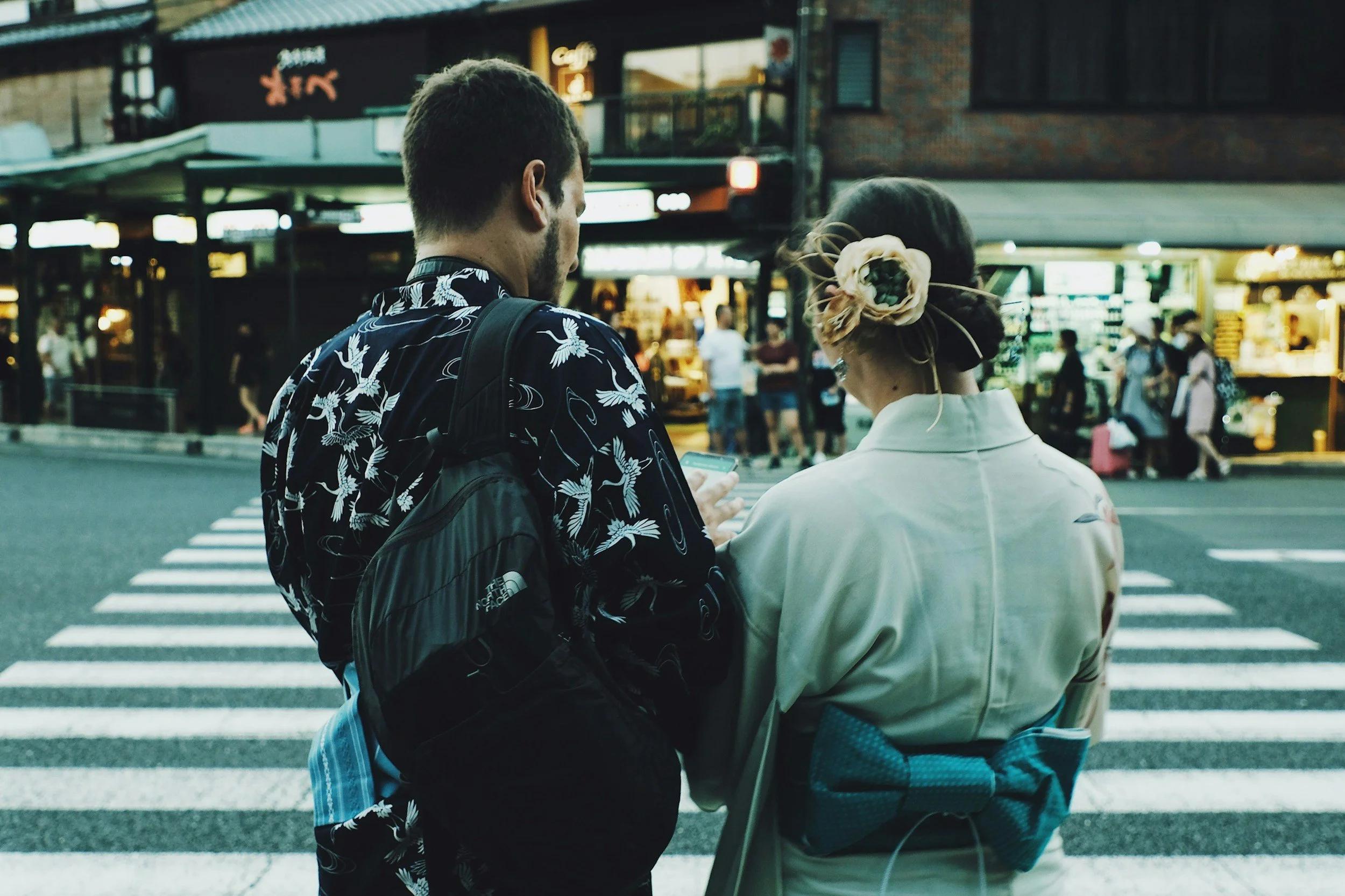 Two people, a man and a woman, standing at a crosswalk in an urban area during evening. The man has short hair, a beard, and is wearing a patterned shirt with a backpack. The woman has dark hair styled with an elaborate flower hair accessory, and is wearing a white or light-colored blouse. They are looking at a smartphone together, with shops and people in the background.