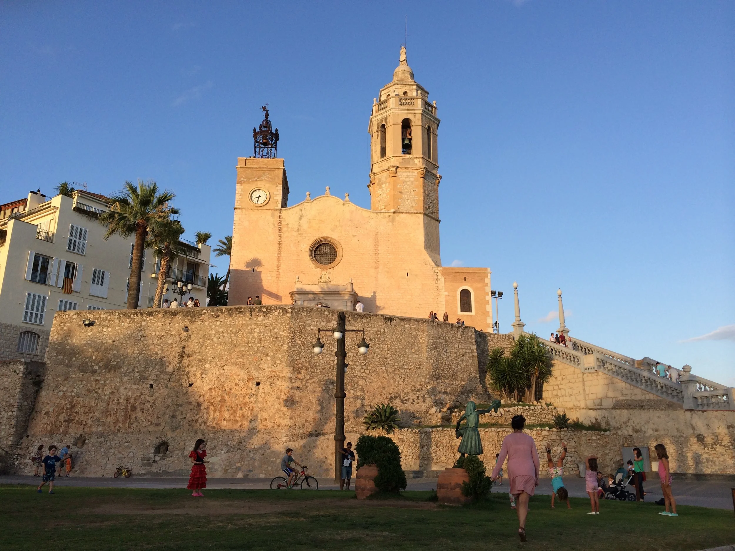 People walking and children playing near a historic church with two towers under a clear blue sky.