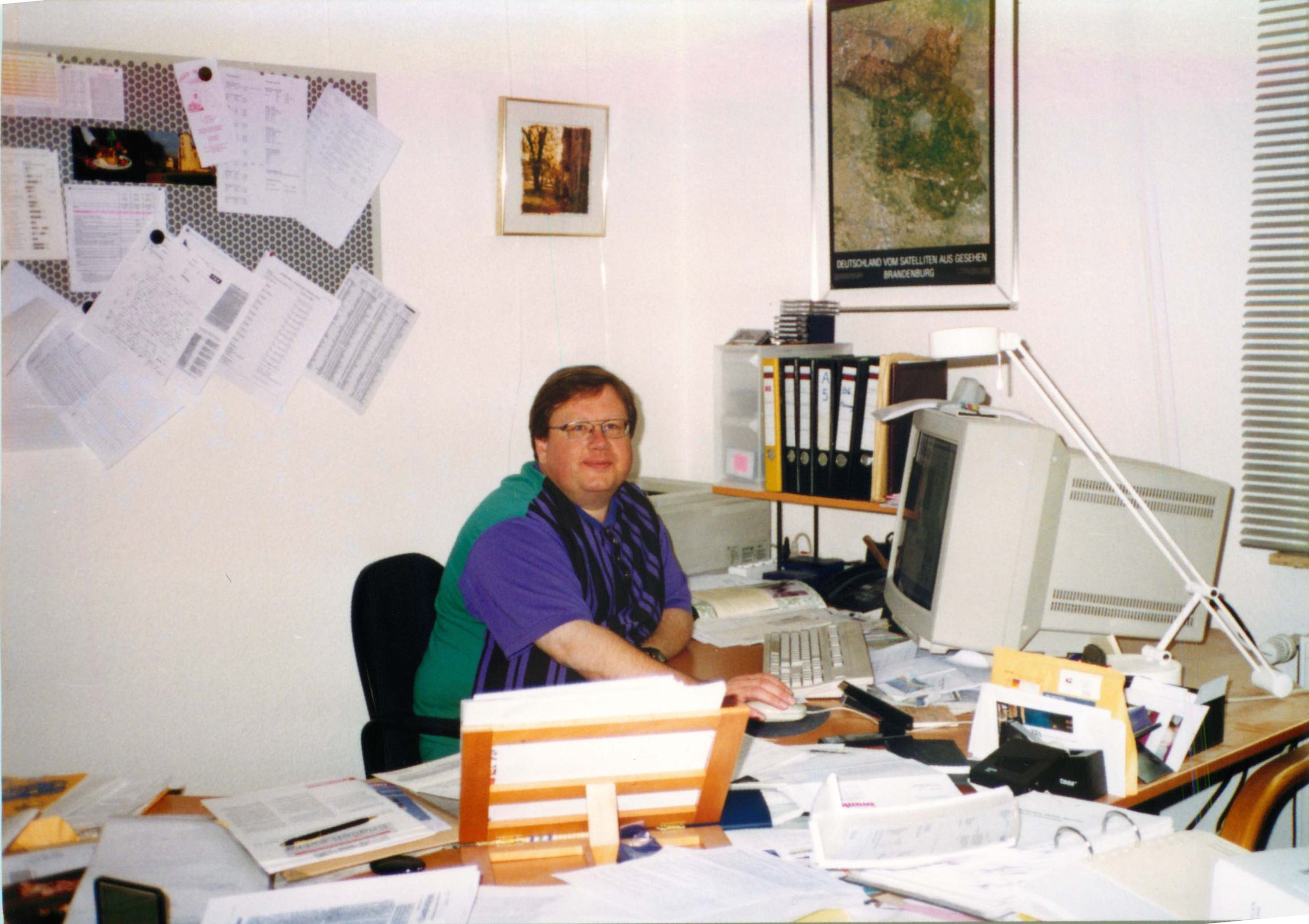 A man with glasses sitting at a cluttered desk with a computer monitor, surrounded by papers, files, and office supplies in a busy office environment.