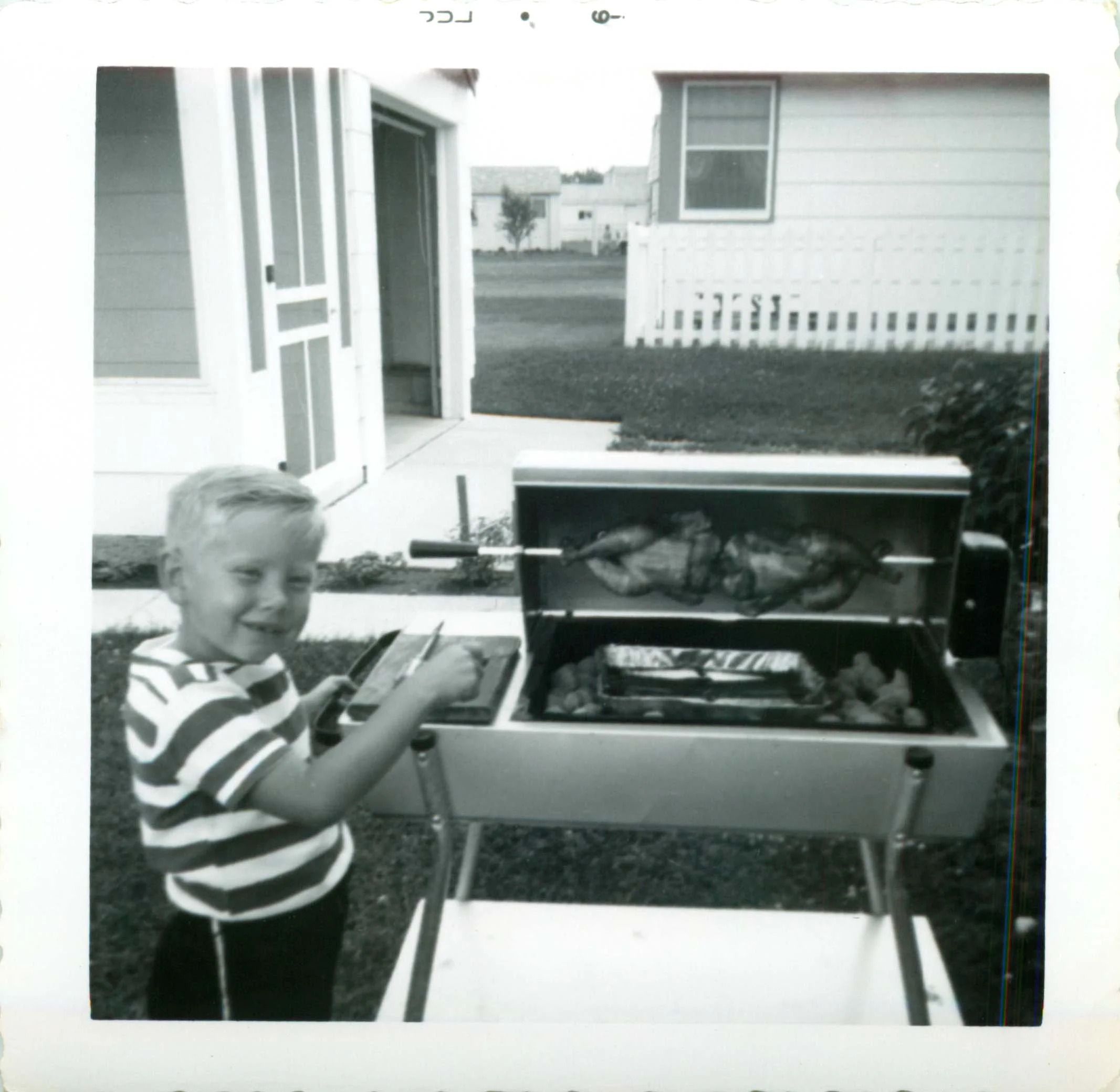 A black and white photo of a young boy with short hair wearing a striped shirt. He is tending a grill, smiling at the camera, with a house and a white picket fence in the background.