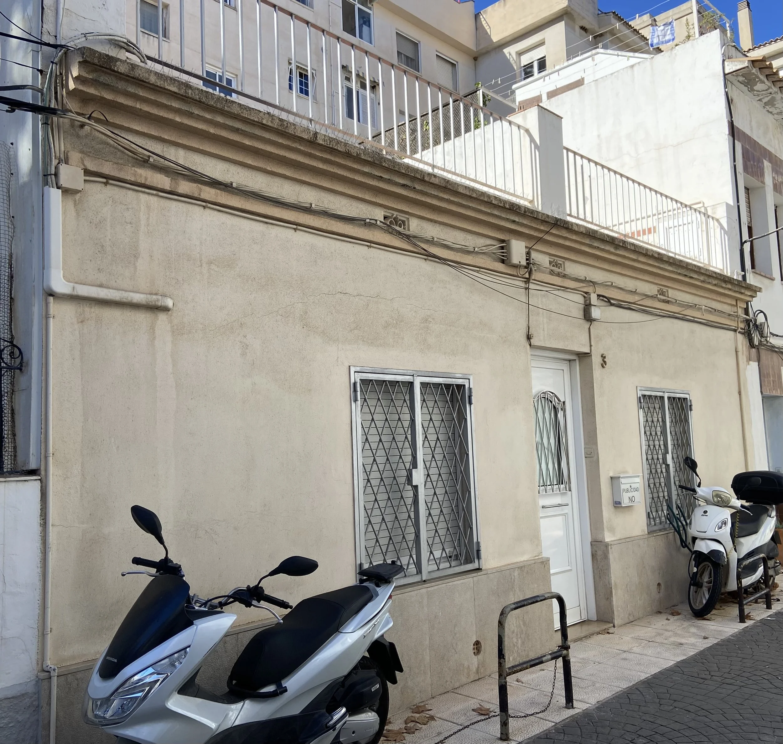 Street view showing a building with metal window bars, scooters parked on the sidewalk, and a white wall with electrical wiring.
