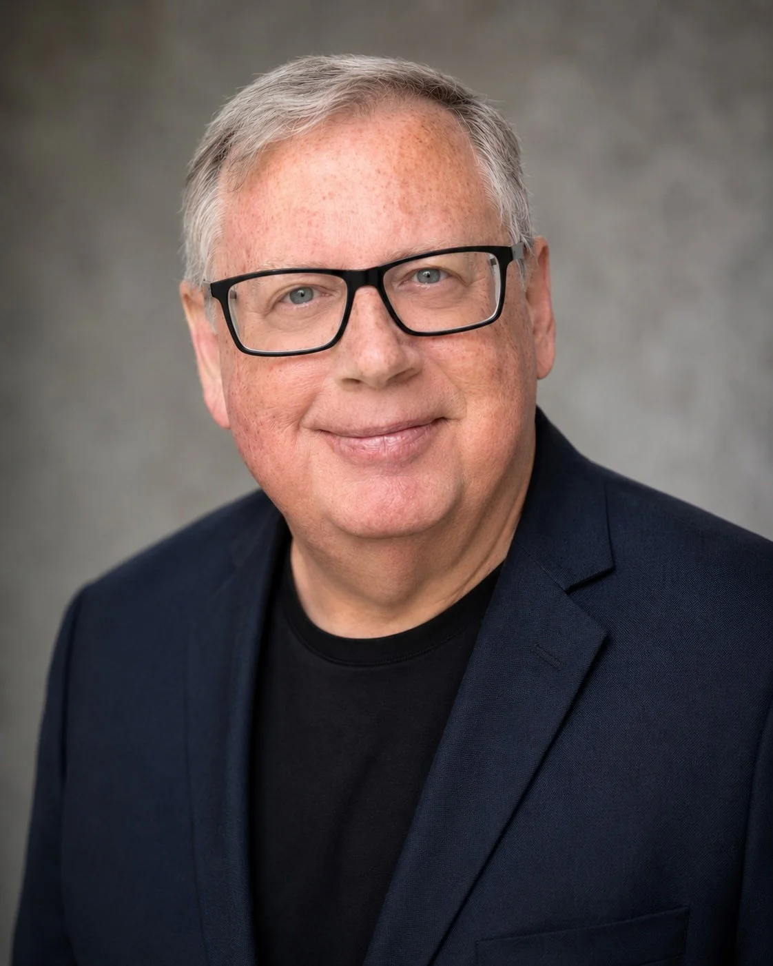 Headshot of a middle-aged man with gray hair, glasses, wearing a dark blazer and black shirt, smiling against a gray background.