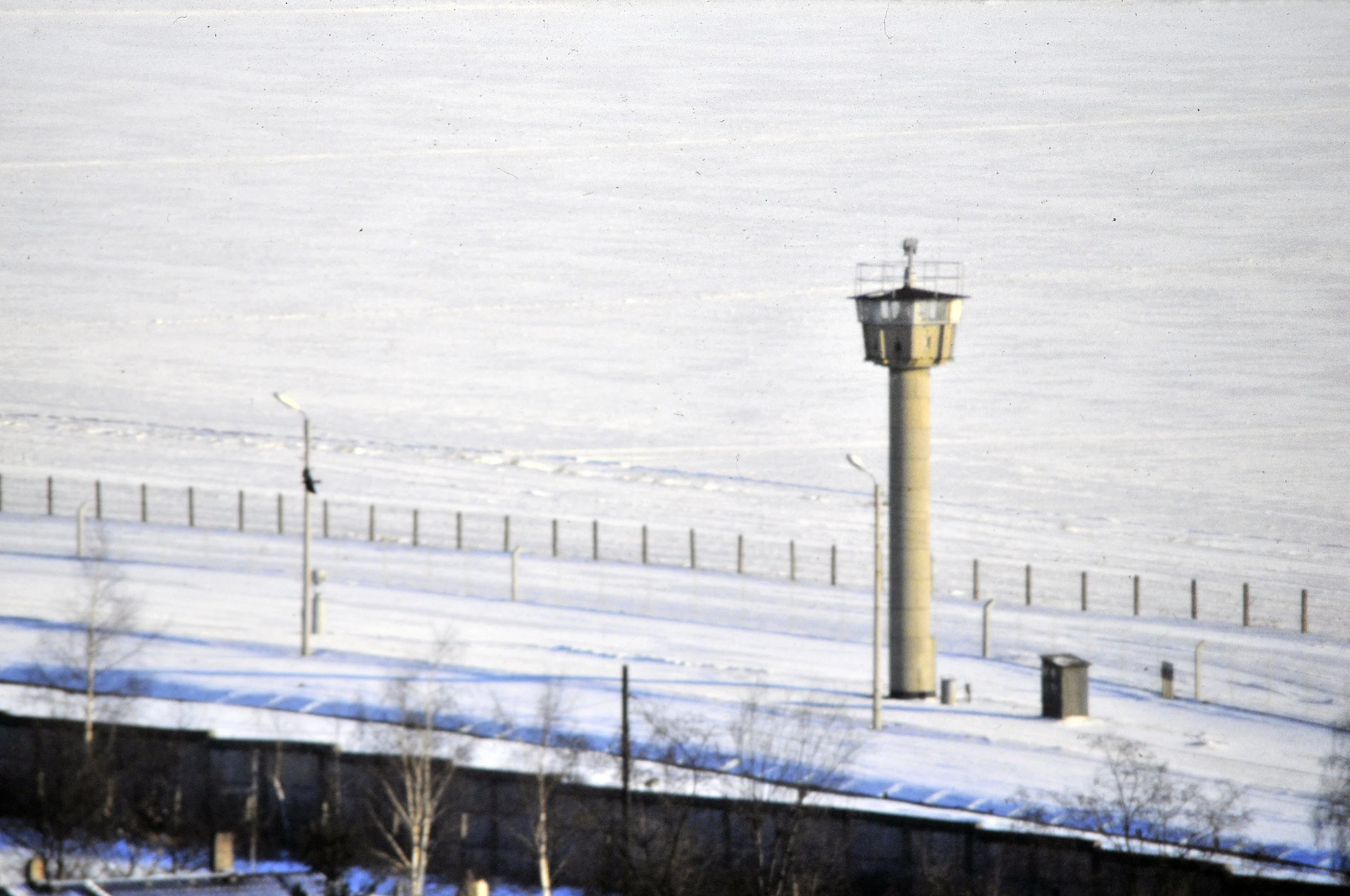 A snow-covered landscape with a tall guard tower, with lines of fences to either side