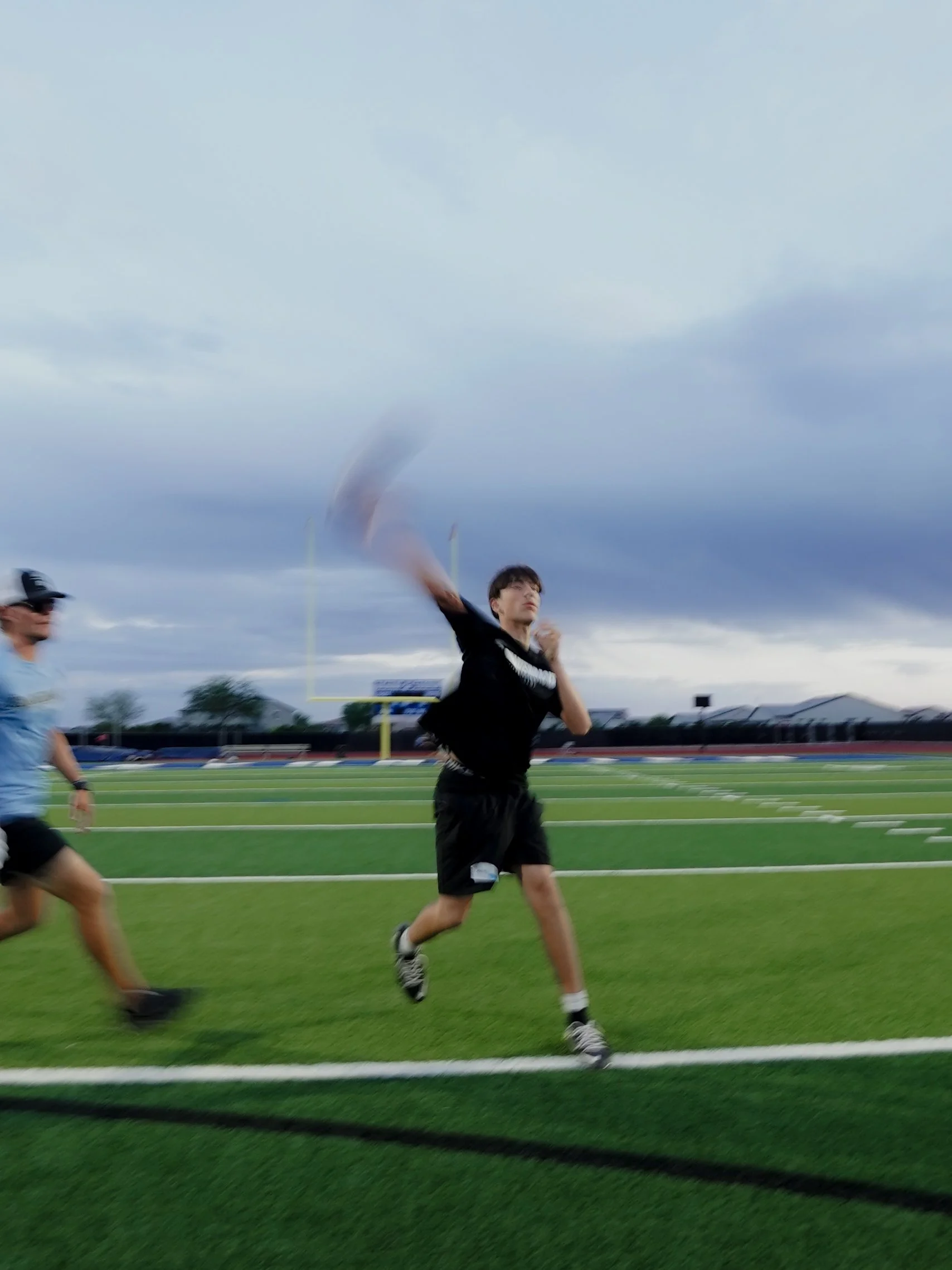 Two young men running on a football field during a track and field event under cloudy skies.